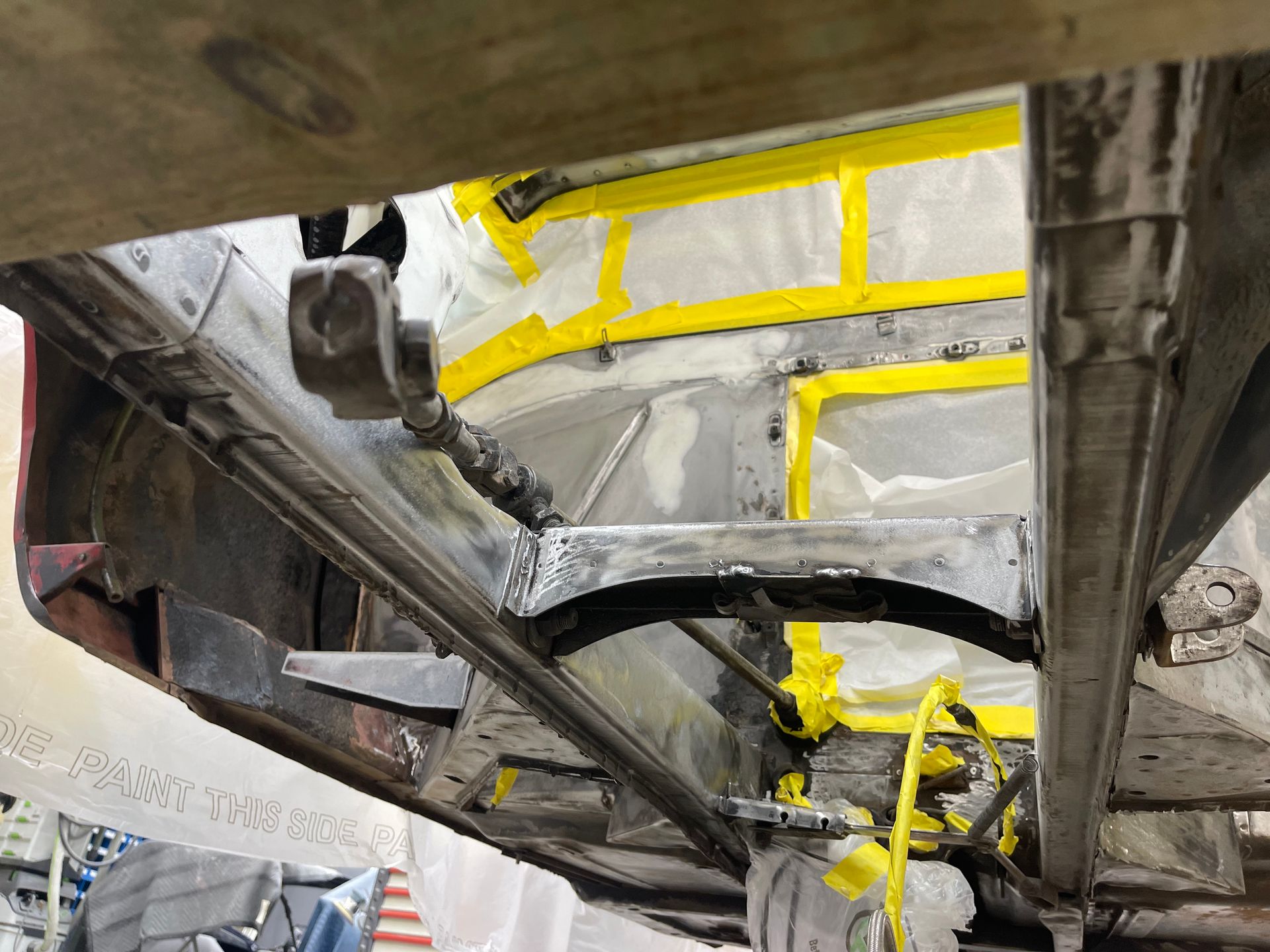 Underside of a vehicle frame during repair, showing metal chassis rails, a support crossmember, and taped-off body sections.