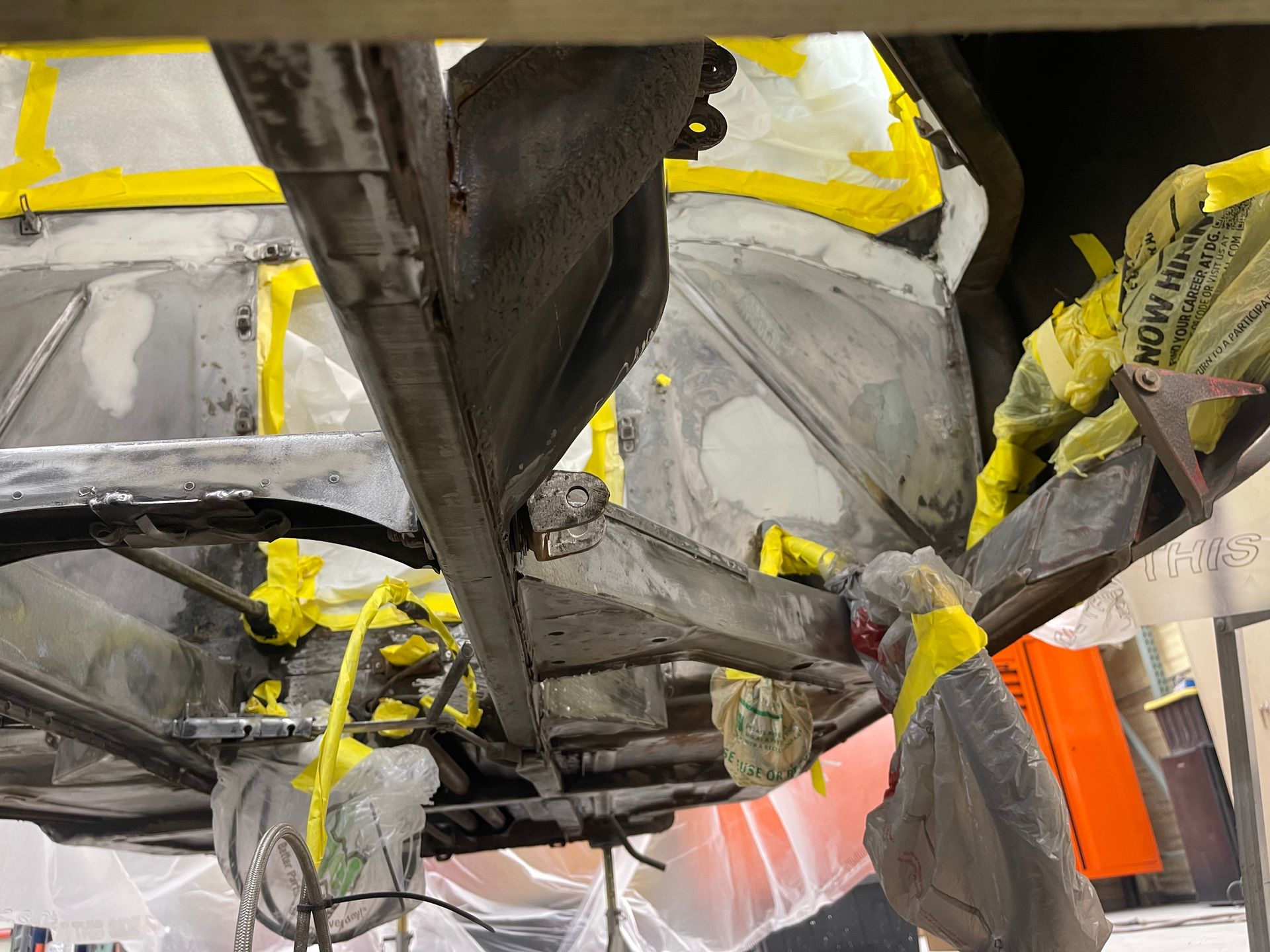 Underside of a vehicle under restoration, showing metal frame components masked with yellow tape and plastic sheeting.