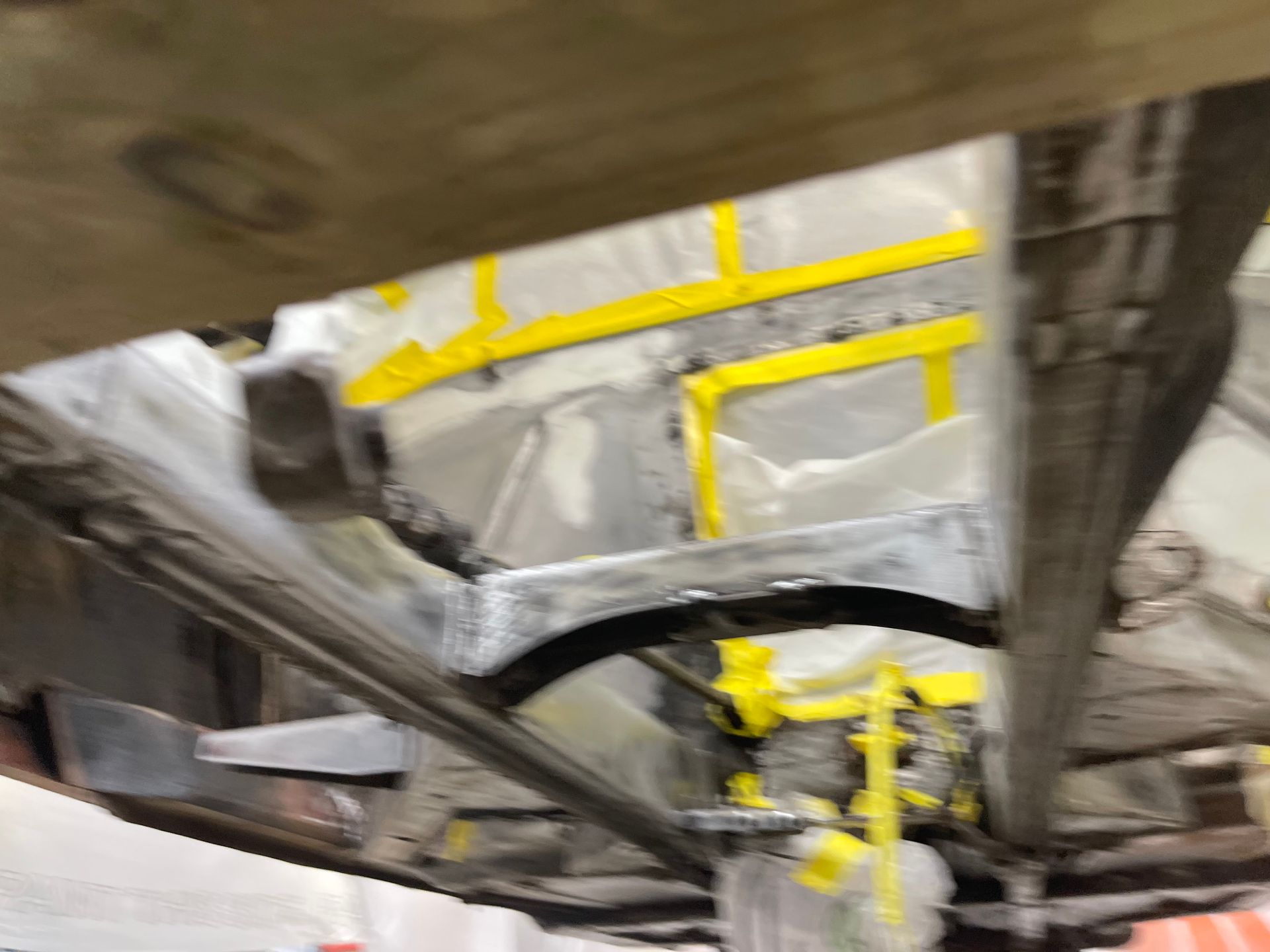 A car frame undercarriage undergoing restoration, featuring exposed metal, welded components, and yellow masking tape.
