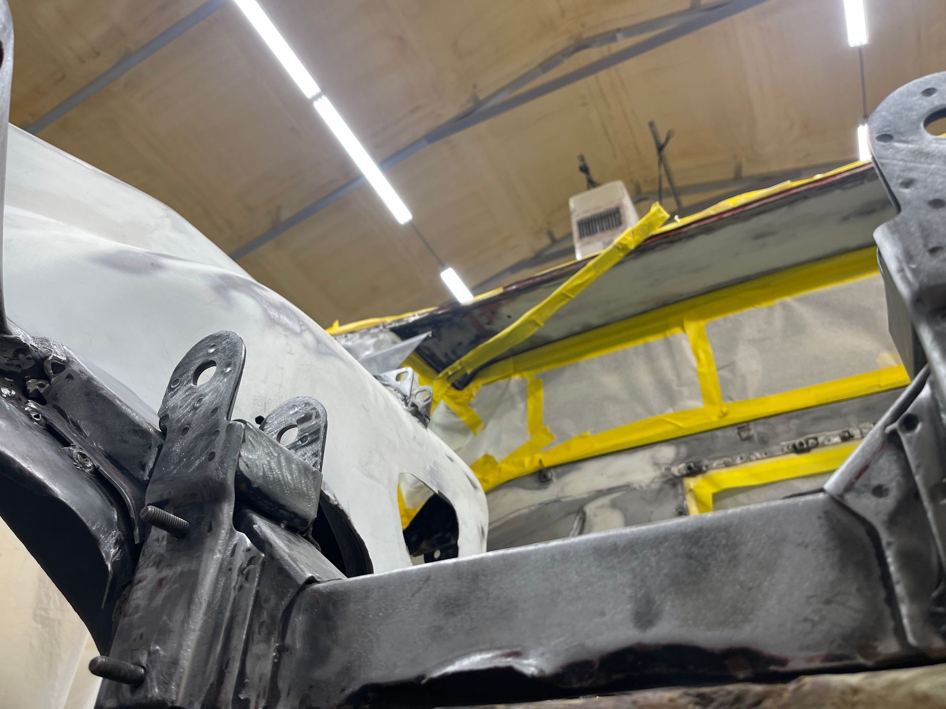 A close-up view of a metal automotive frame under repair, with visible welding and yellow masking tape in a workshop.