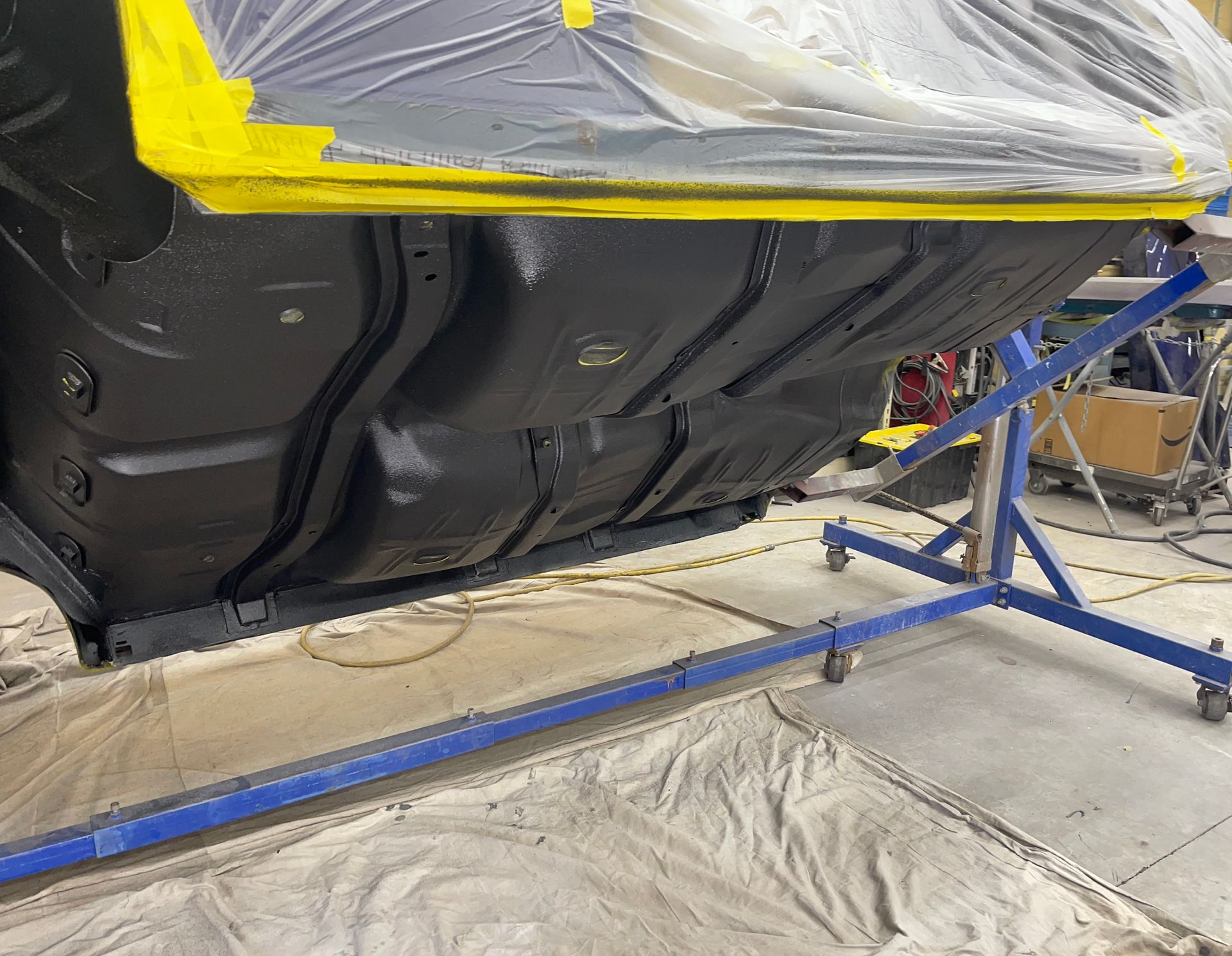 A car chassis on a rotisserie stand in a workshop, freshly painted black with taped-off areas covered in clear plastic.