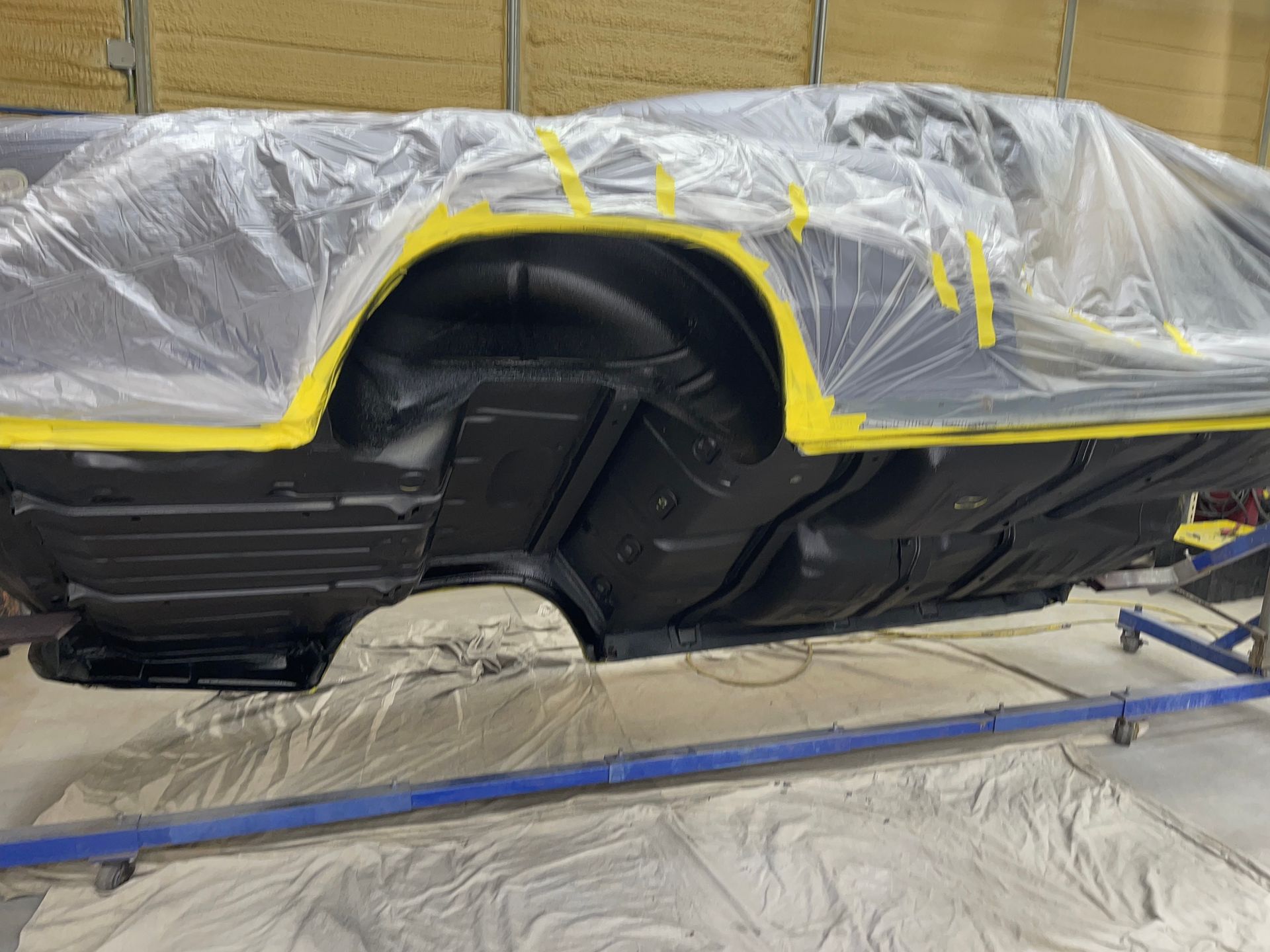A car body on a rotisserie, painted matte black with yellow masking tape along the wheel wells and covered in plastic.