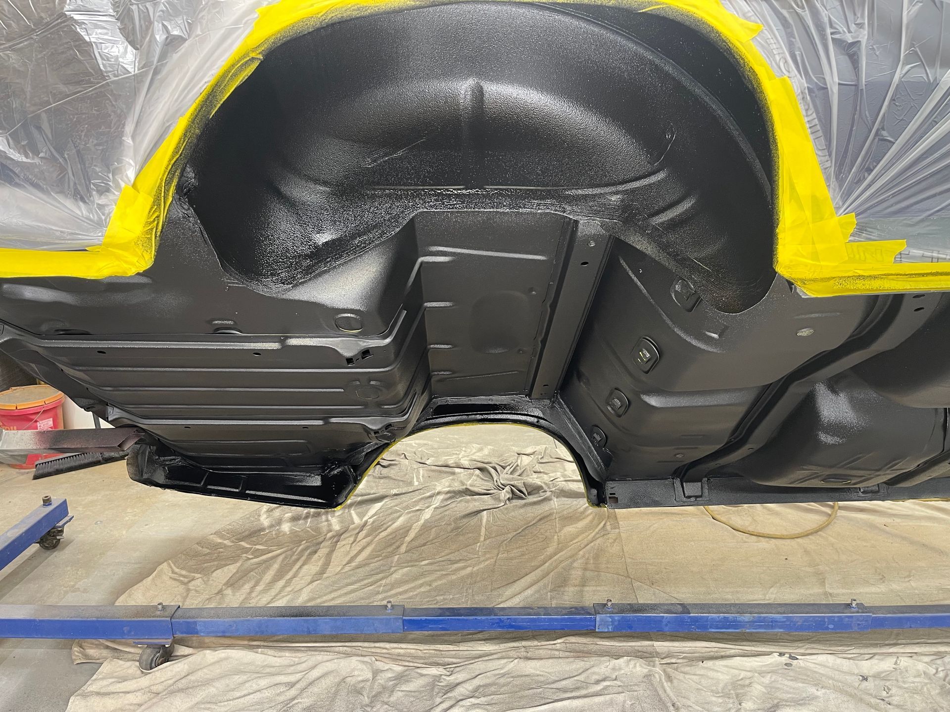 A vehicle wheel well and undercarriage freshly coated in black protective paint, with yellow masking tape on the edges.