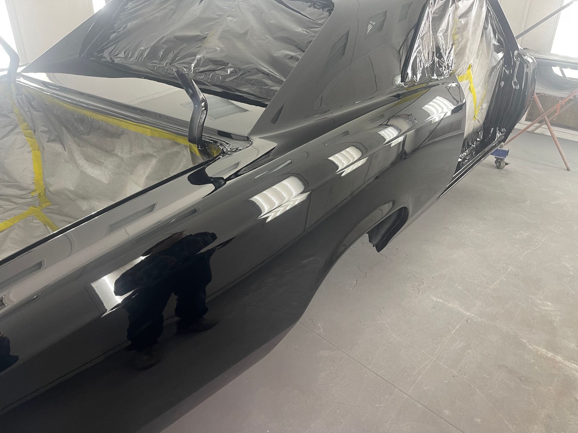 A glossy black car body in a spray booth, partially covered with plastic and masking tape for a custom paint job.