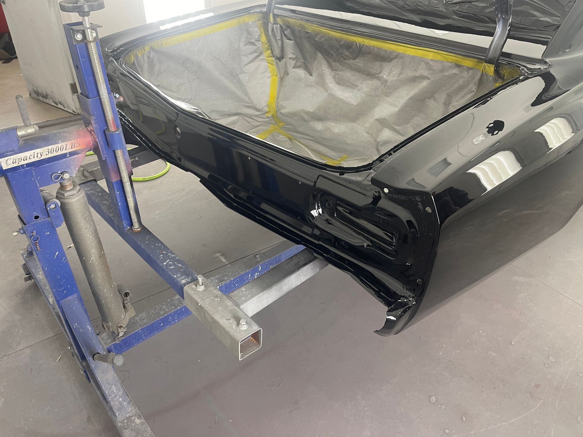 The rear section of a glossy black car body mounted on a blue industrial stand in a workshop.