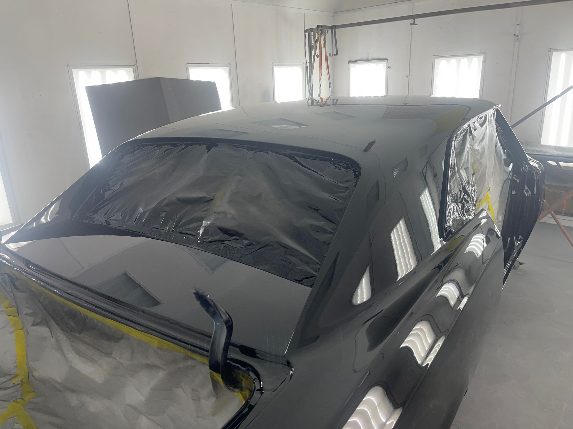 A black car, masked with plastic sheeting, sits in a paint booth during a professional restoration process.