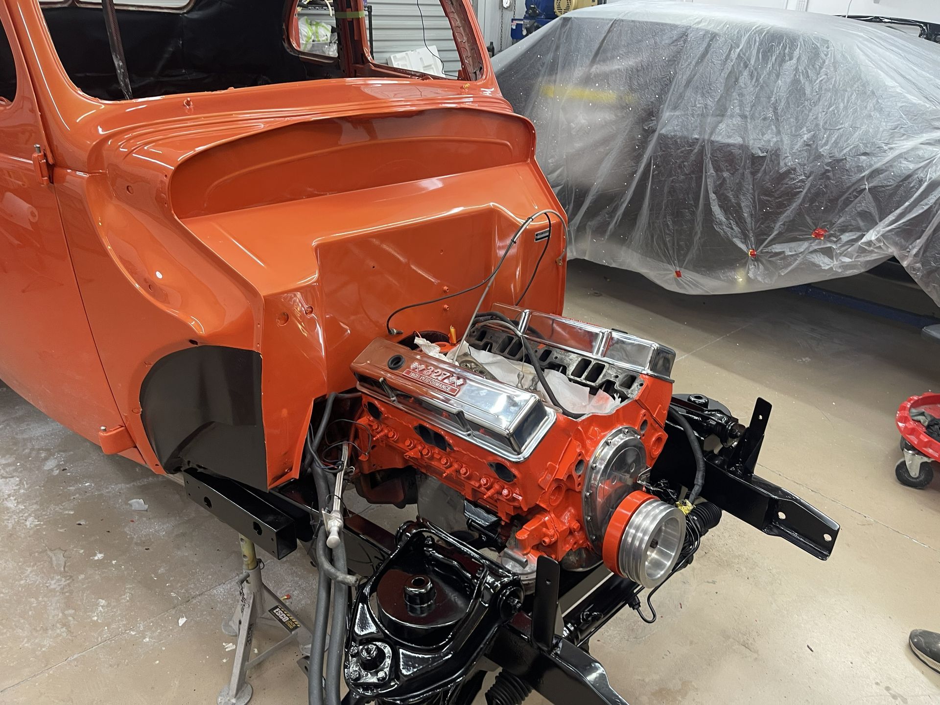 An orange vehicle chassis with an exposed V8 engine in a garage, next to a car covered in plastic sheeting.