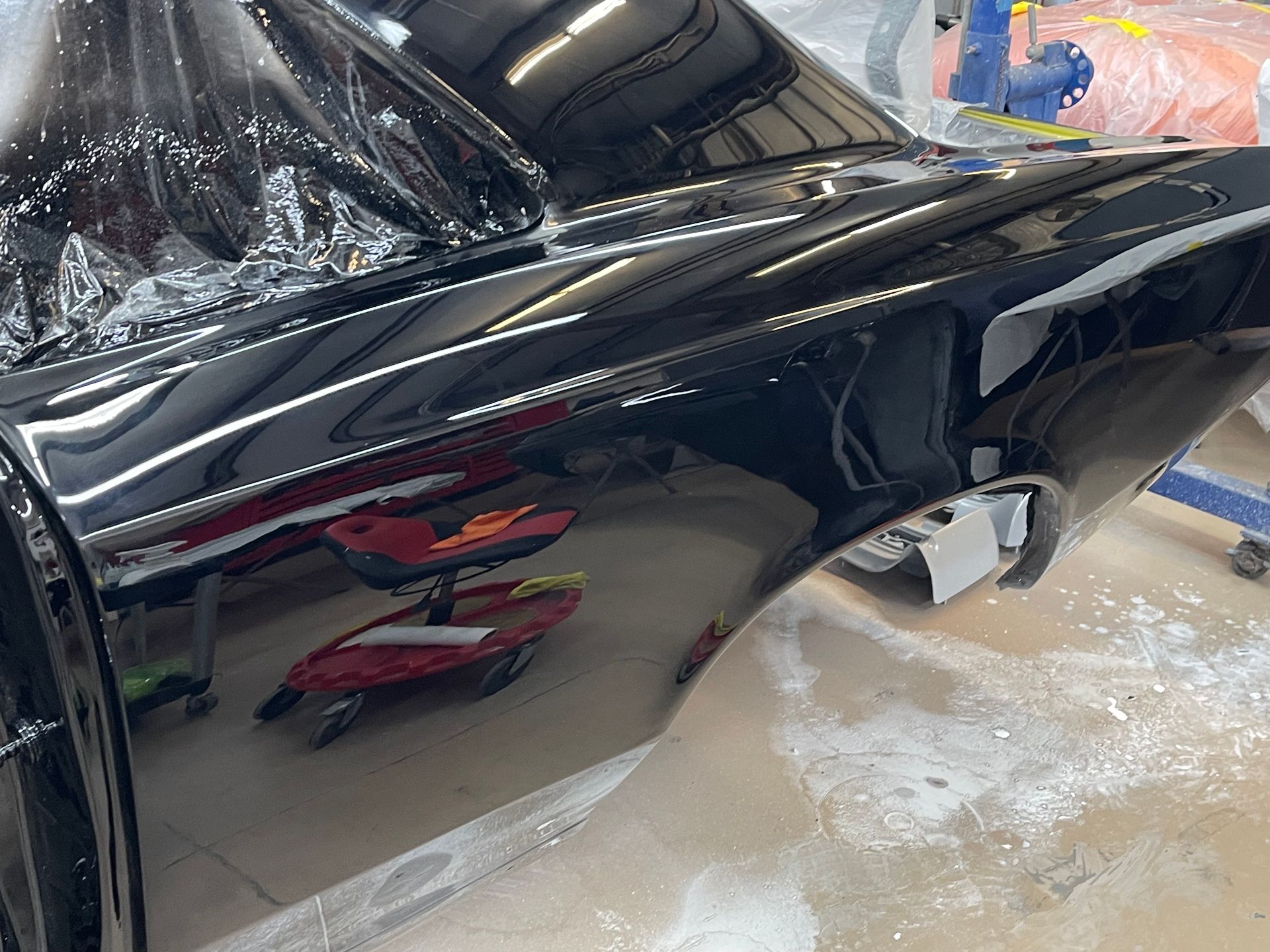 A freshly painted, glossy black car panel sits in an auto body shop, reflecting the surrounding workspace.