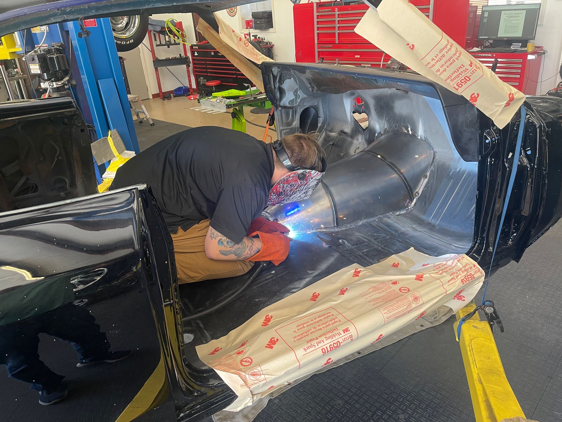 A person wearing protective gear uses a welding torch on the interior metal frame of a black car in a workshop.