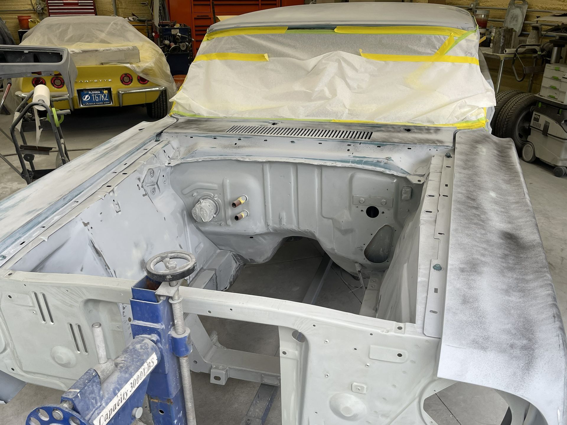 A vehicle undergoing auto body restoration in a shop, with a masked windshield and an empty, primed engine bay.