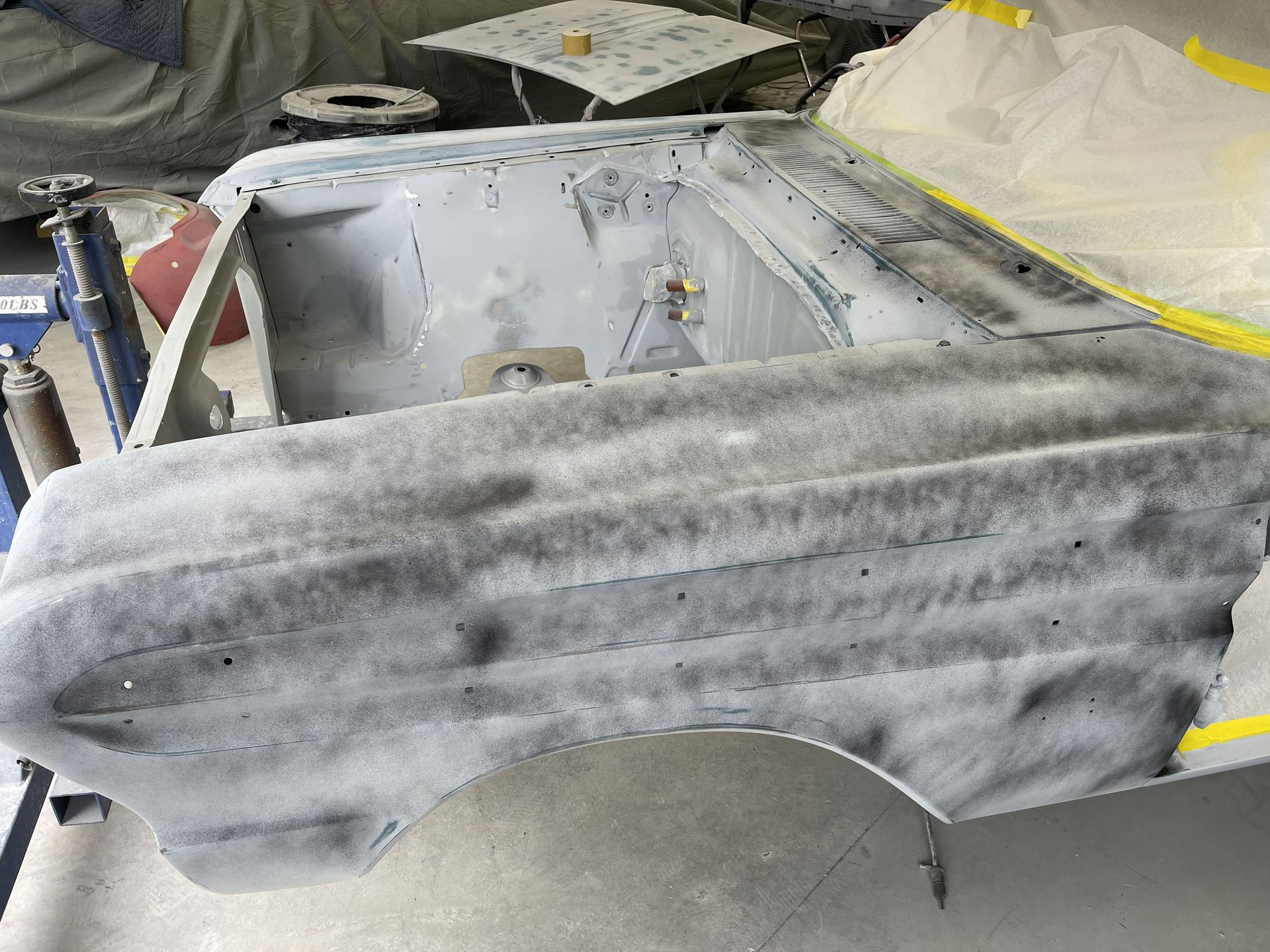 A car's engine bay and front fender in a garage, covered in grey guide coat spray during automotive body restoration.