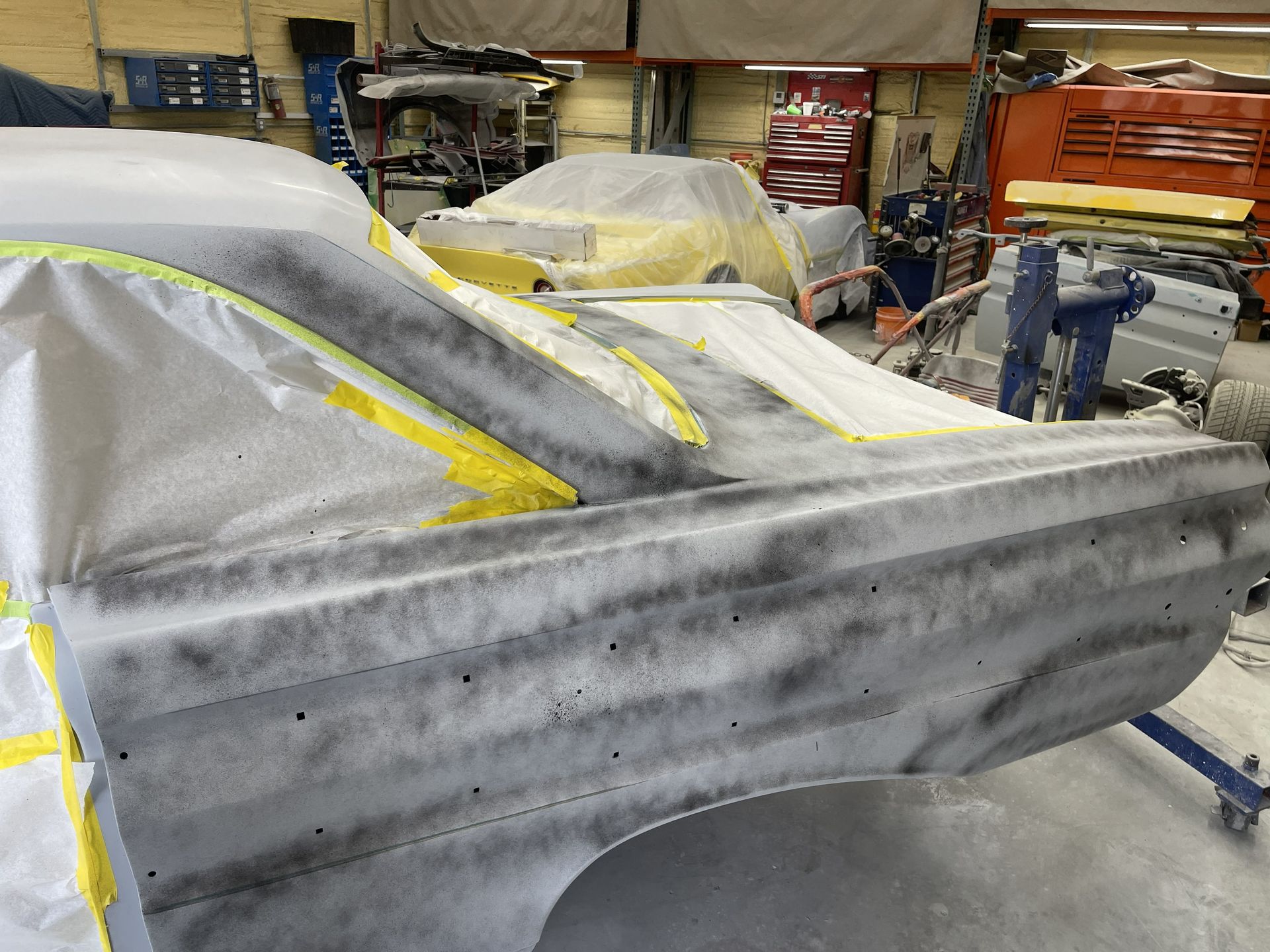 The rear side of a car body is coated in gray primer and masked with yellow tape inside an auto body repair shop.