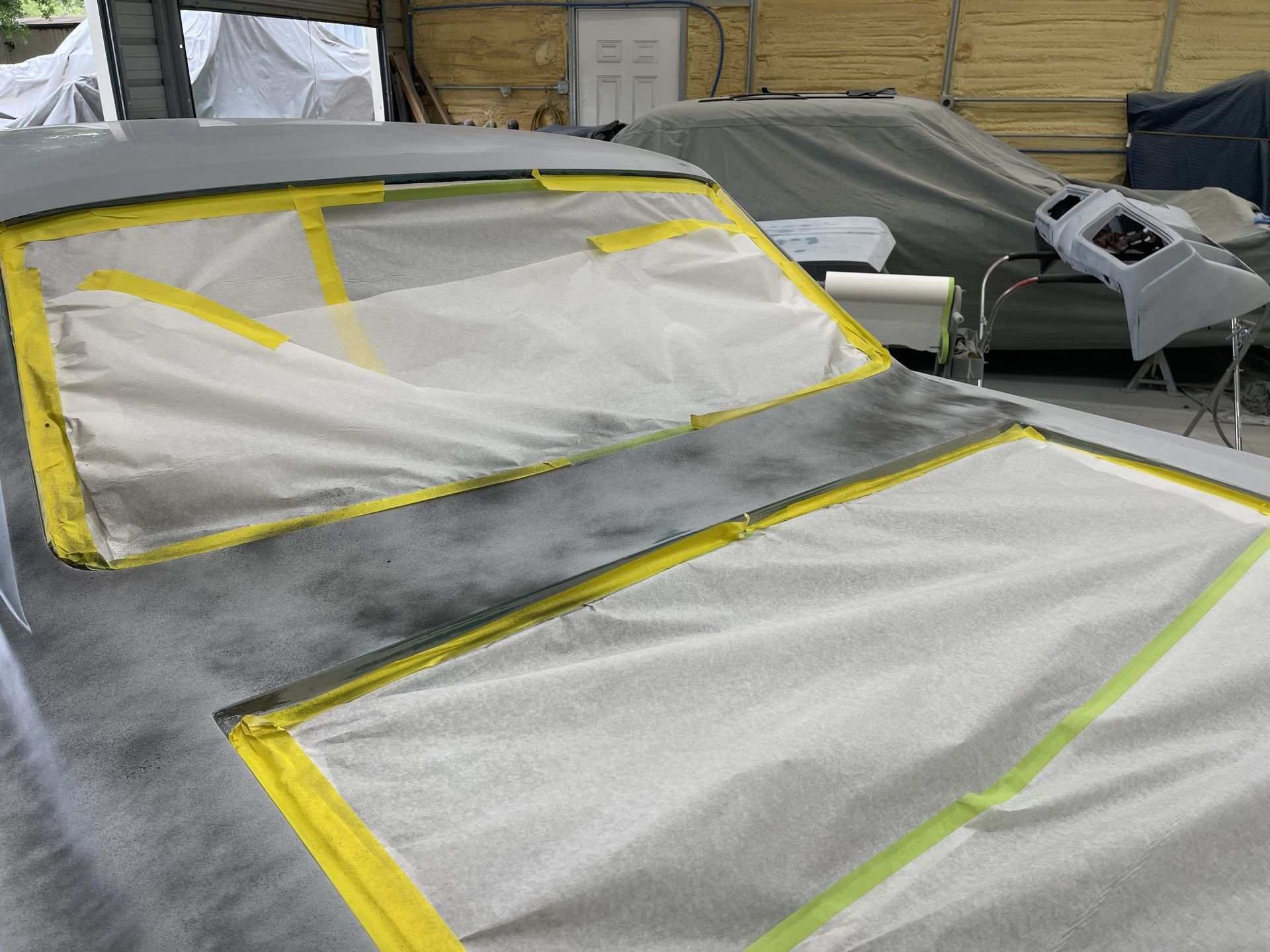 A car body being prepared for painting, with windows and hood masked off with white paper and yellow painter's tape.