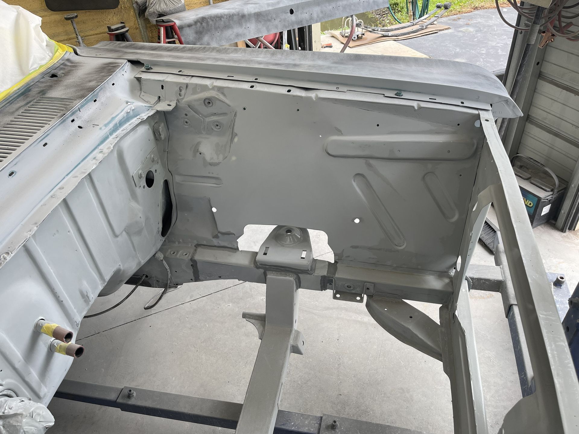 The engine bay of a car under restoration, painted in light gray primer, showing the firewall and front frame rails.