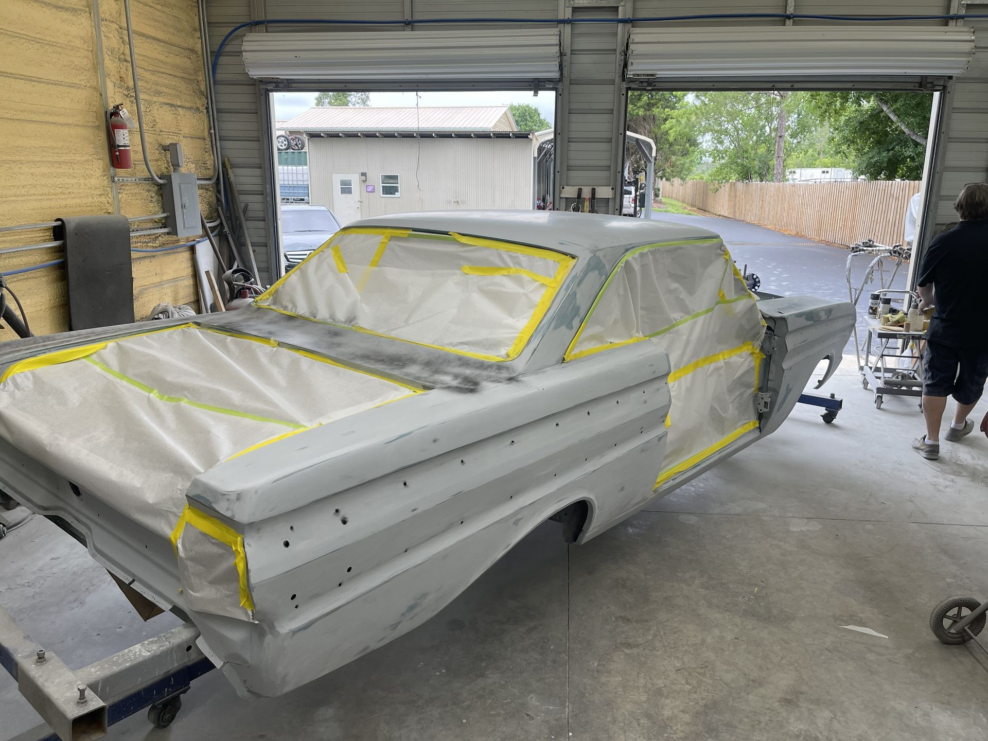 A grey car body in a workshop, masked with tape and paper for painting, with a person standing nearby.