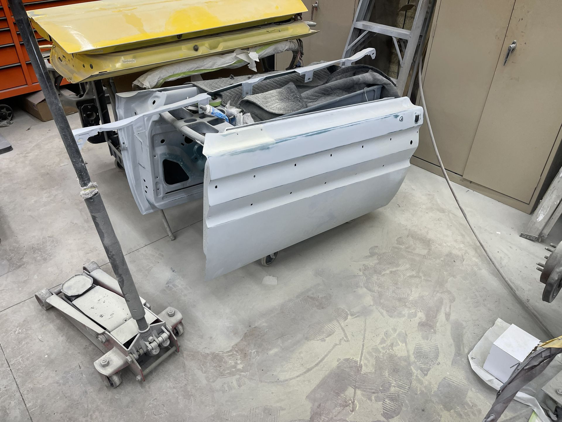 A primed white car door stands in a garage, next to a yellow door panel and a hydraulic floor jack.