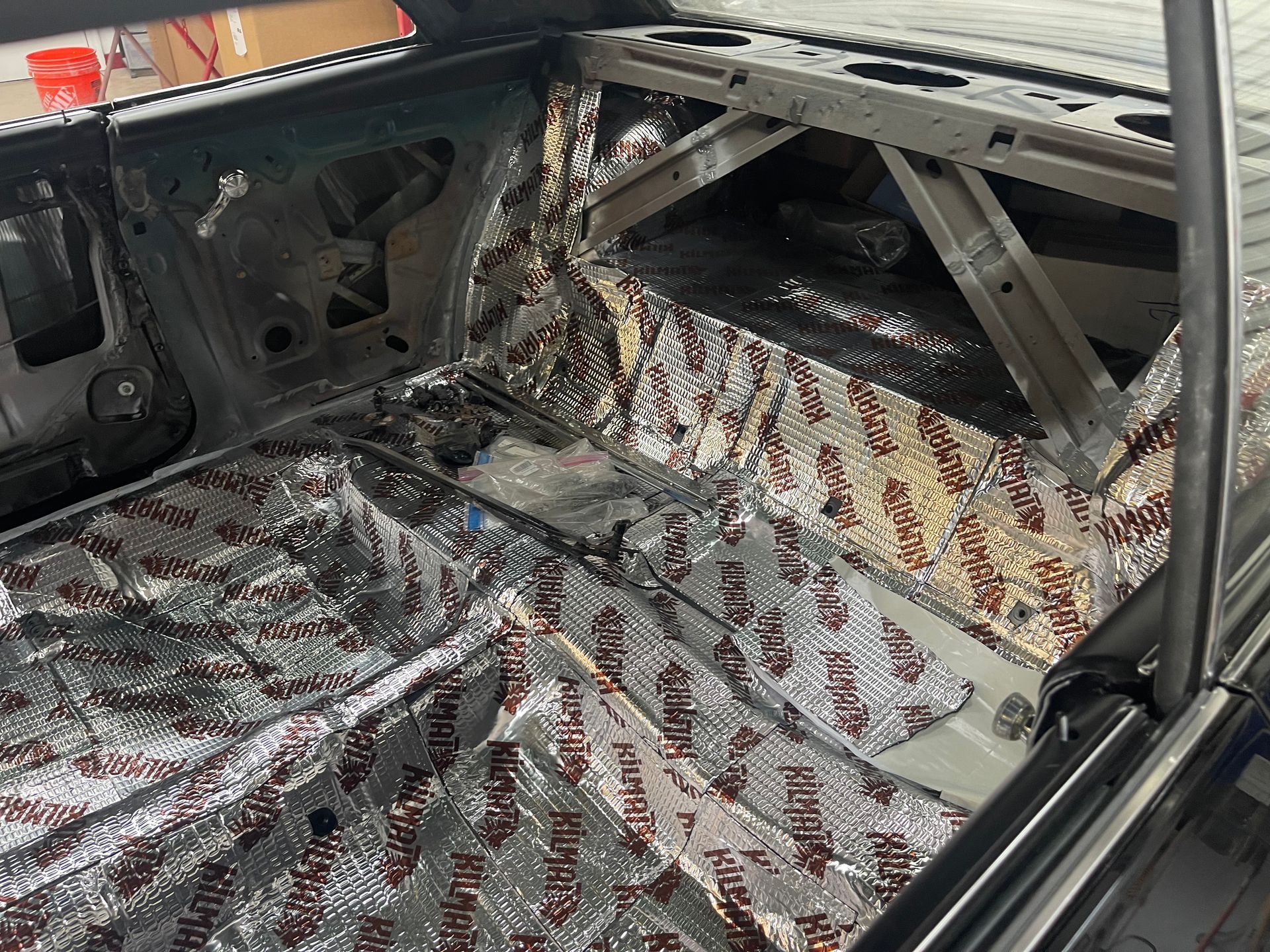 The stripped interior of a car floor and rear shelf covered in silver sound-deadening insulation material.