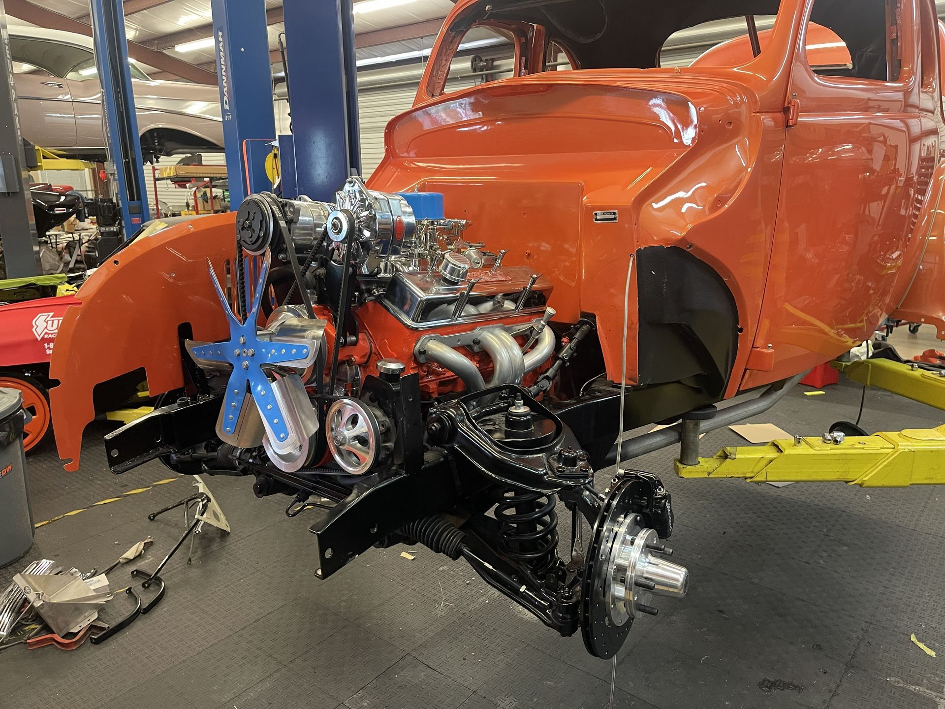 A bright orange vintage car chassis undergoing engine work in a brightly lit automotive repair shop.