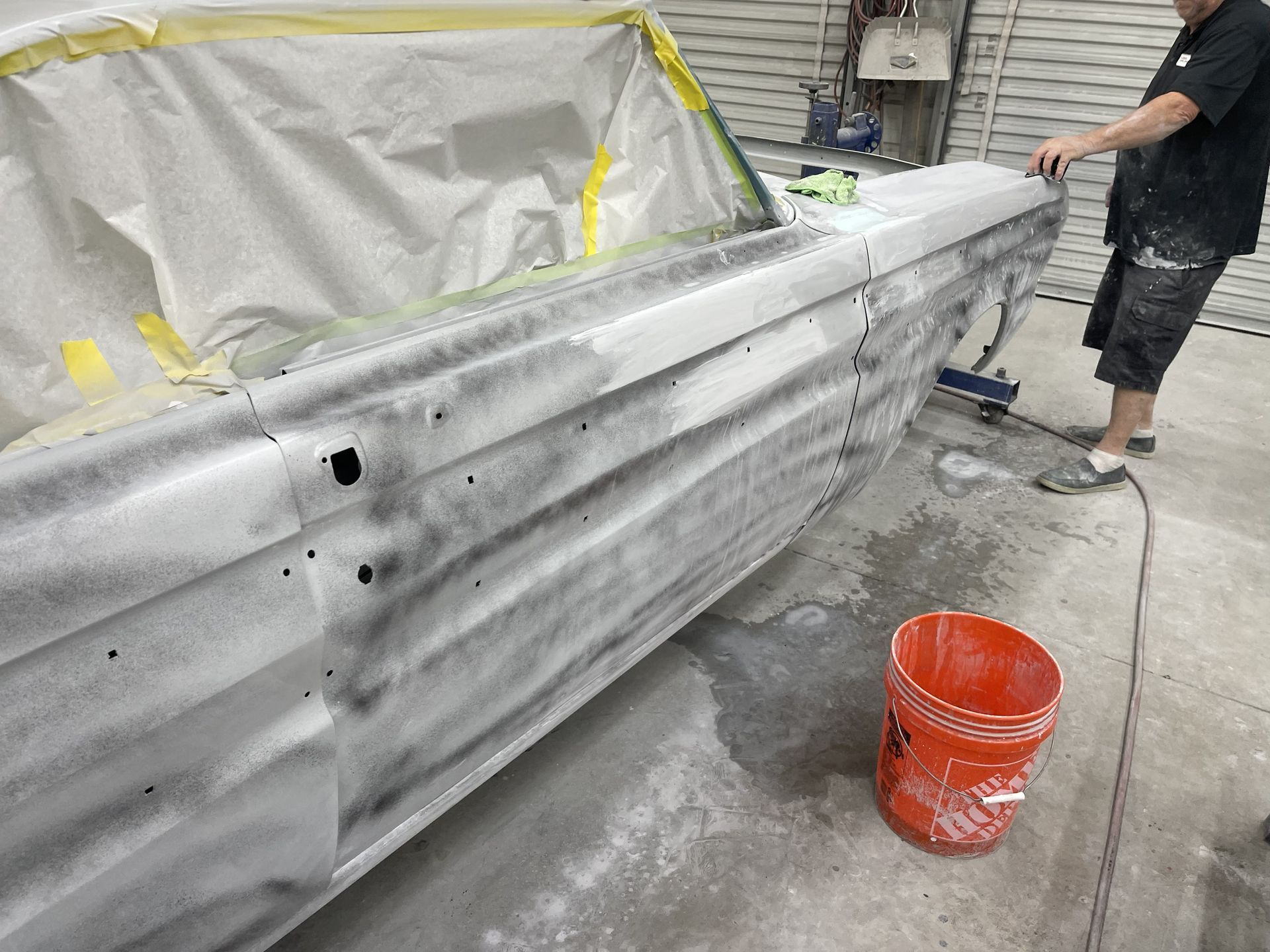 A person sanding the side of a classic car in a workshop, with masking paper on the windows and an orange bucket nearby.