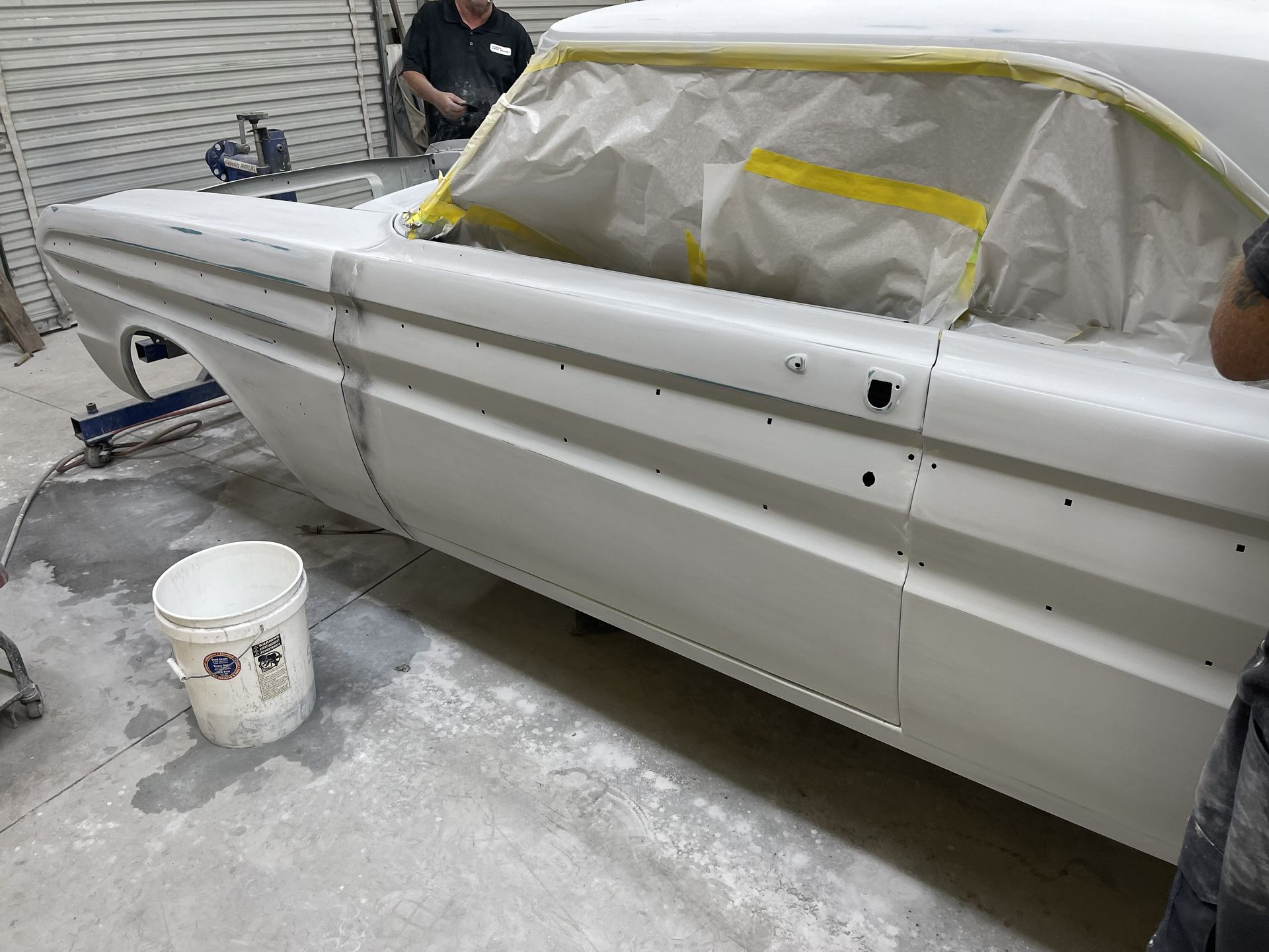 A light-colored, primer-coated classic car body sits in a garage, with taped windows ready for painting.