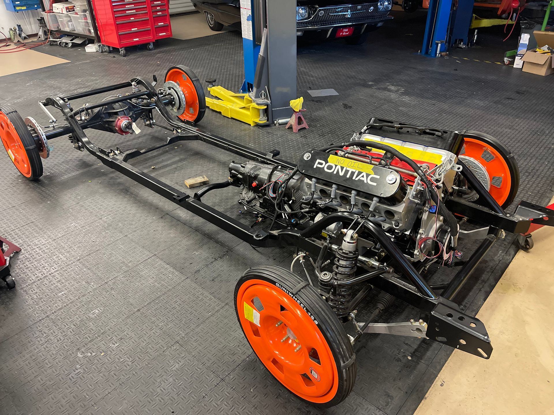 A car chassis with a Pontiac engine and bright orange wheels sits on a garage floor.