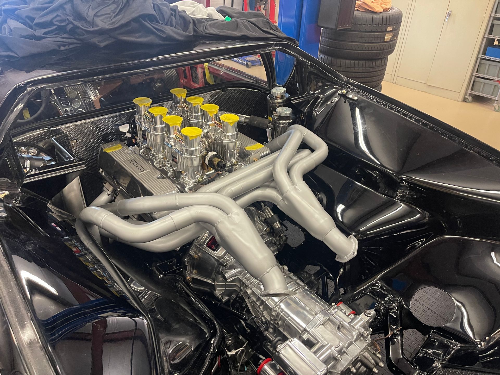 A high-performance V8 engine with silver exhaust headers and yellow-capped velocity stacks inside a black car chassis.