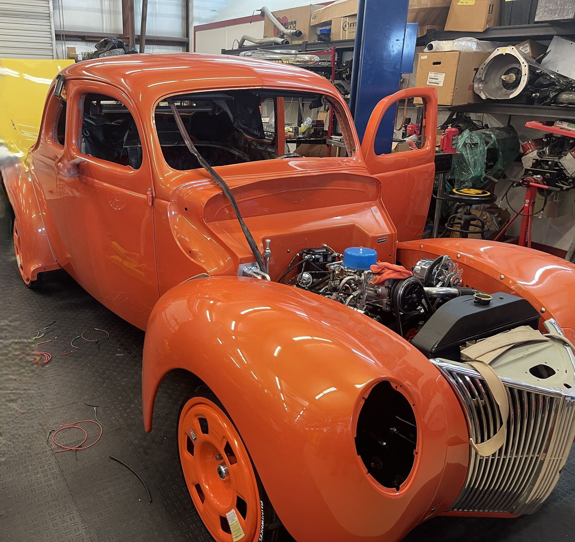 An orange vintage Ford hot rod sits in a workshop with its hood removed and engine visible.
