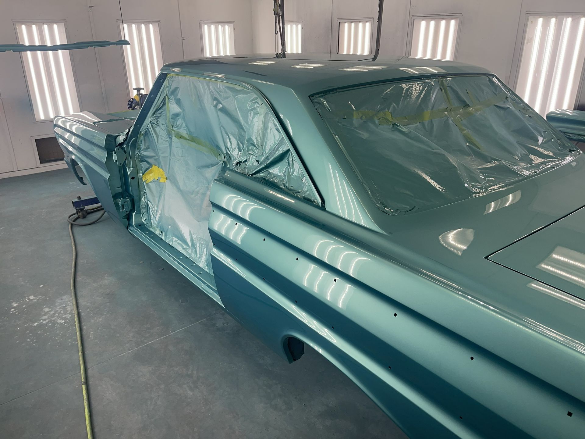 A classic car painted a glossy teal sits in a bright, industrial spray booth, with windows masked by protective plastic.