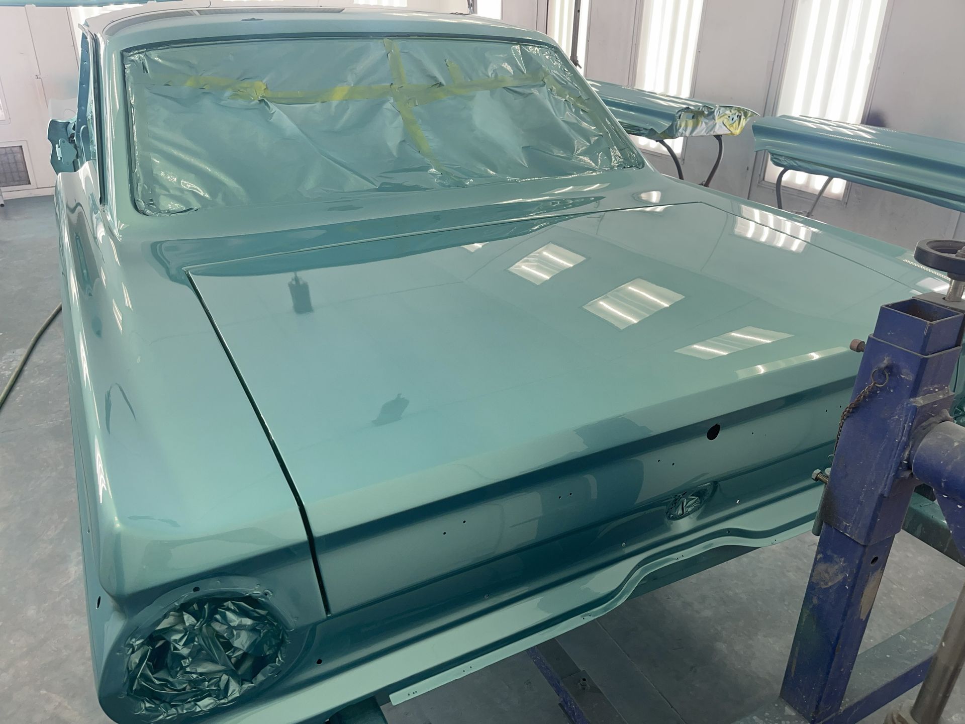 A classic car painted in light blue, freshly coated and covered in masking tape, inside a spray paint booth.