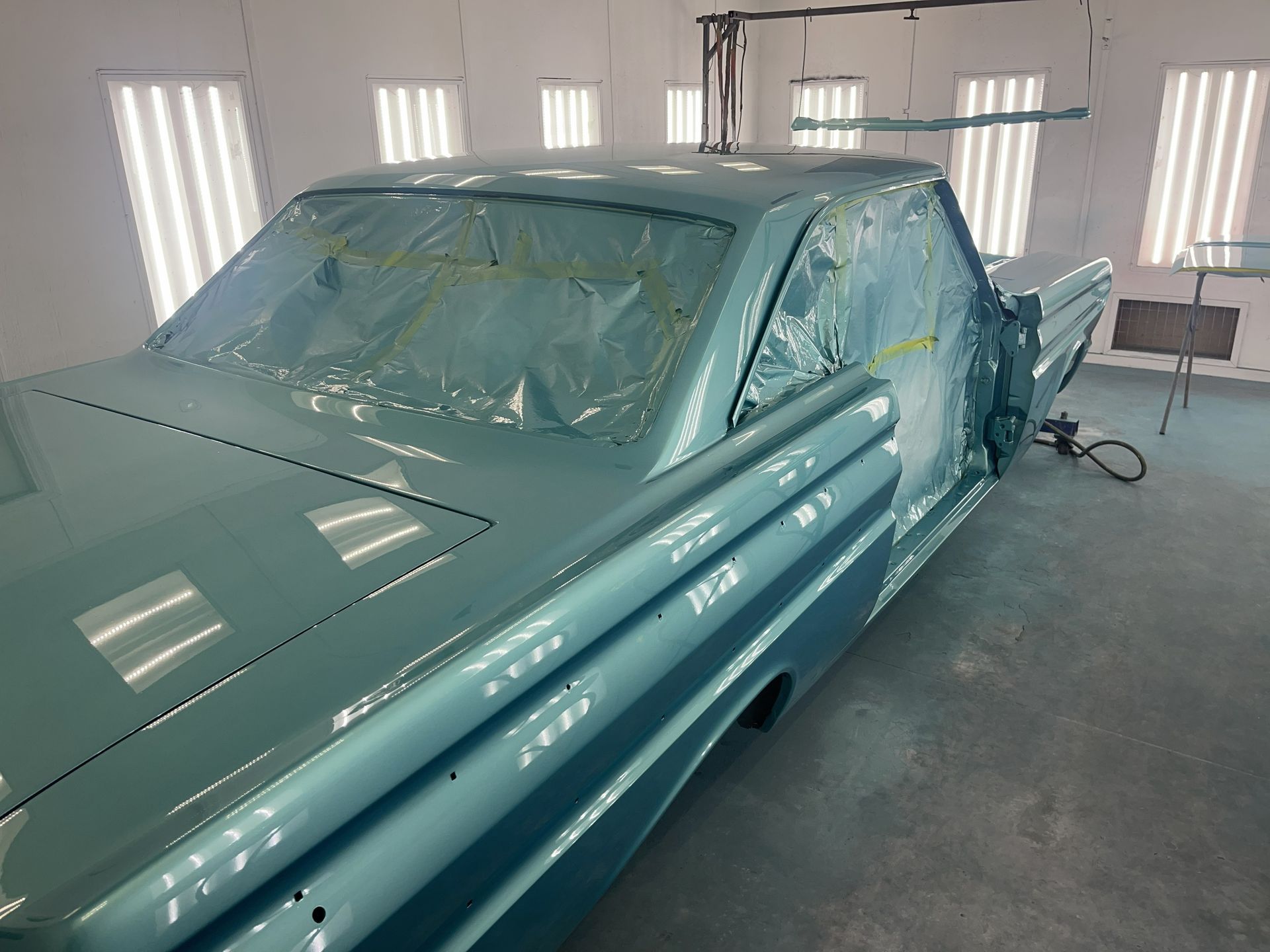 A freshly painted, light blue classic car inside a professional automotive paint booth with plastic masking.