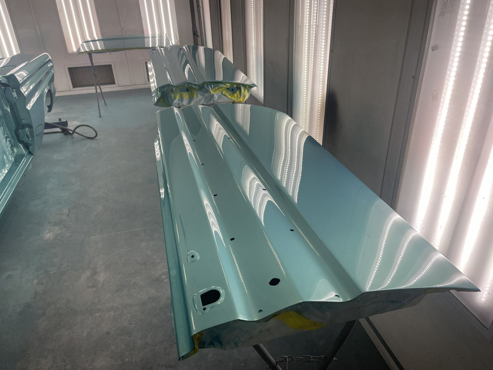 A freshly painted light blue car door panel rests on a stand in an automotive spray booth with bright overhead lighting.