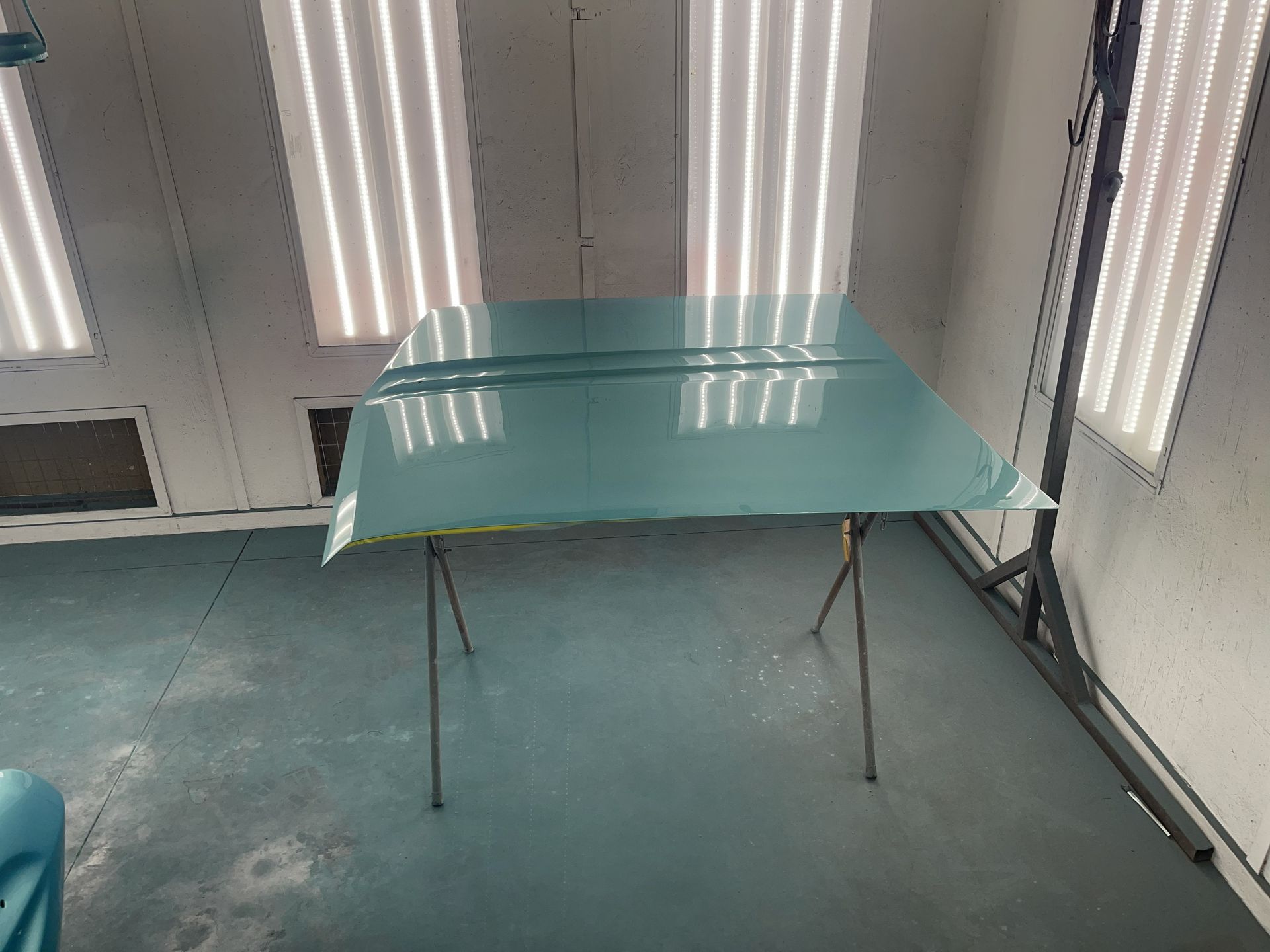 A freshly painted light blue car hood resting on a metal stand inside a brightly lit automotive spray booth.