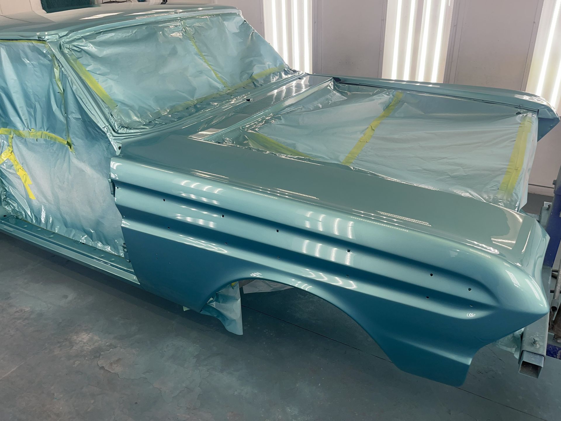 A light blue vintage car body in a workshop, partially covered in protective masking tape and film.