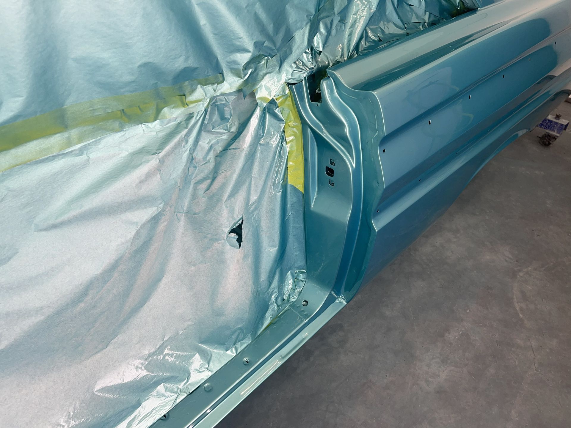 A light blue car door panel being painted, with protective plastic masking the interior frame.