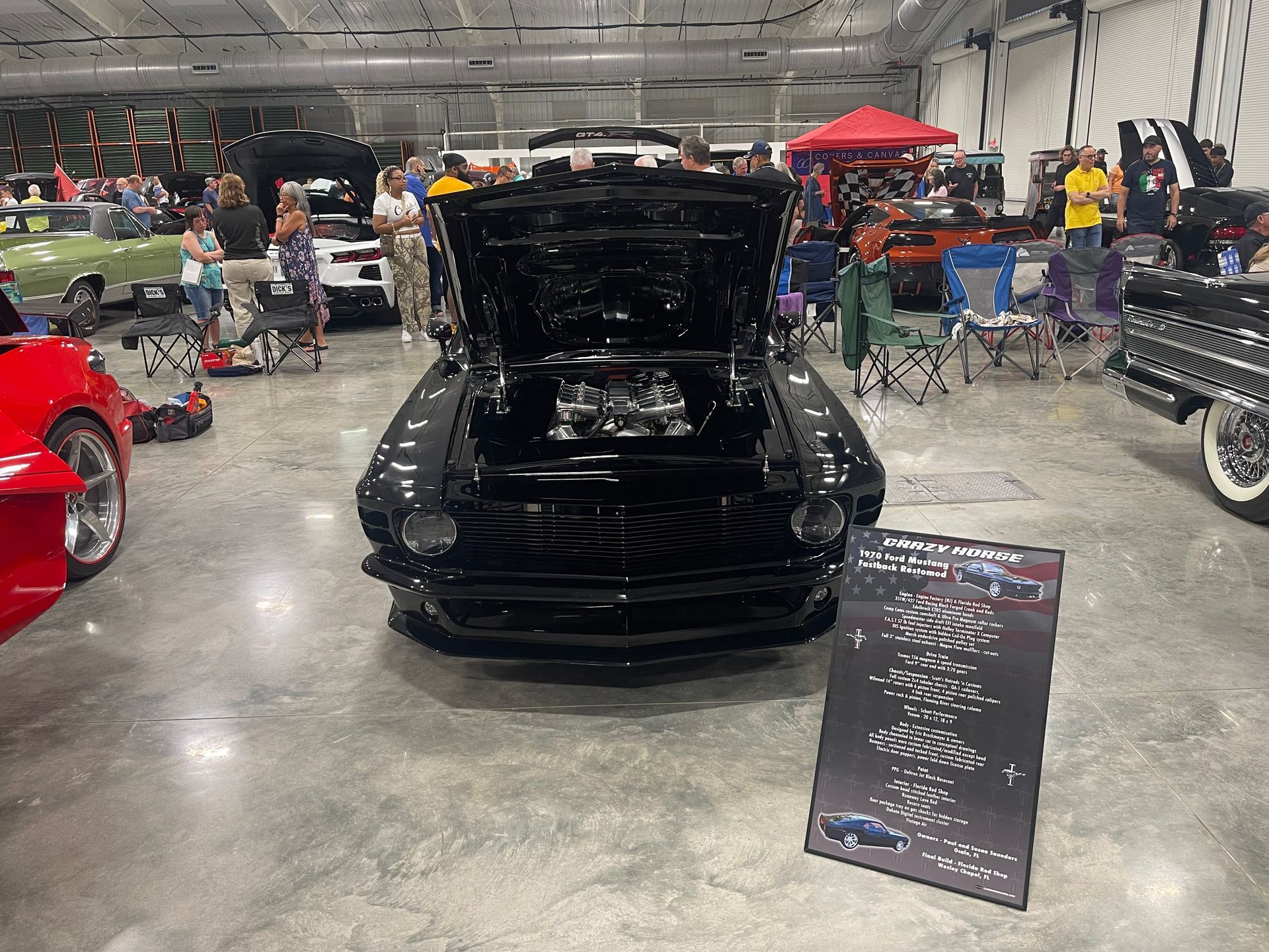 A black vintage muscle car with an open hood on display at an indoor car show, flanked by spectators and other vehicles.