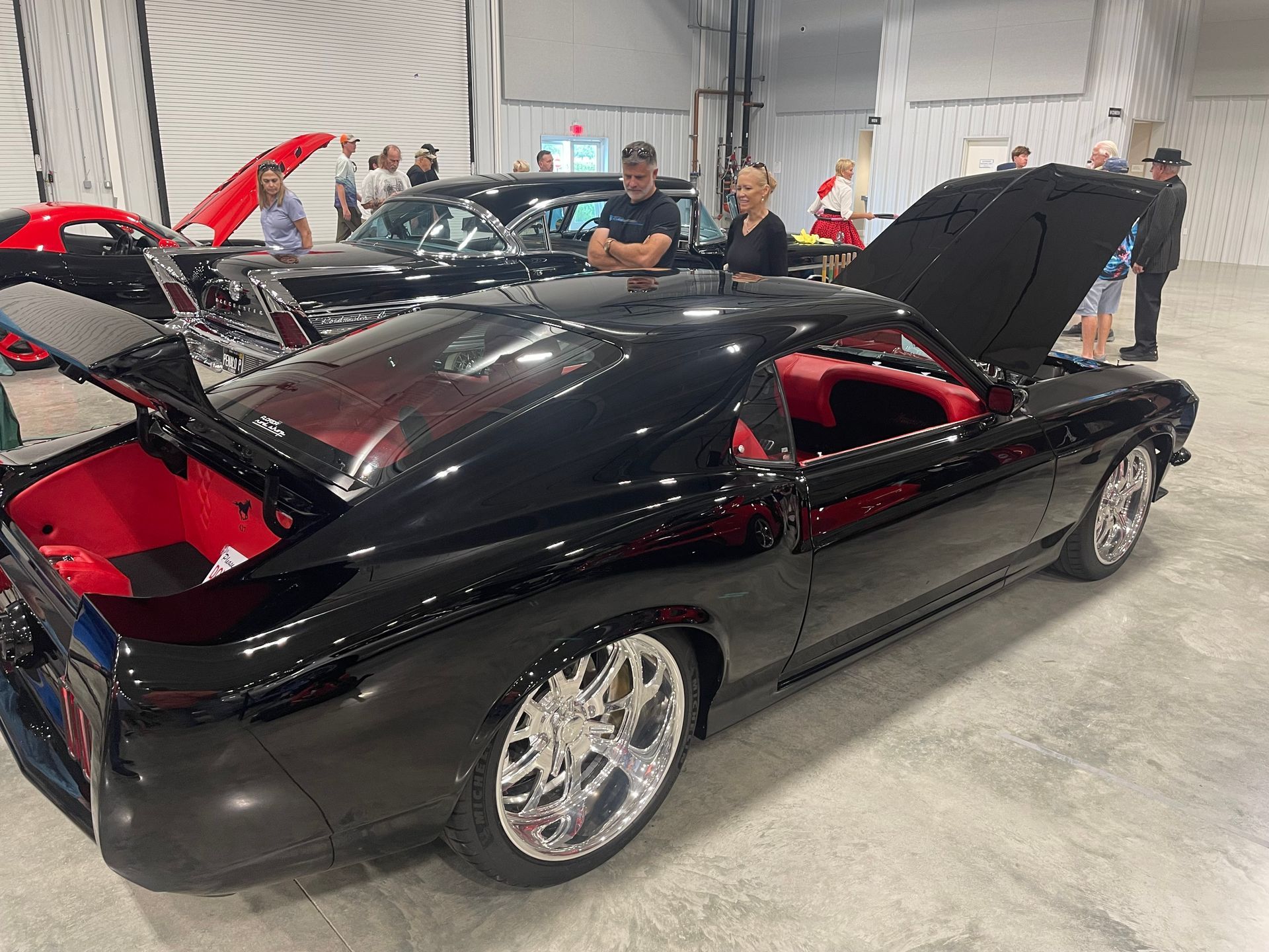 A sleek black classic muscle car with an open hood and trunk is displayed at an indoor car show with people nearby.