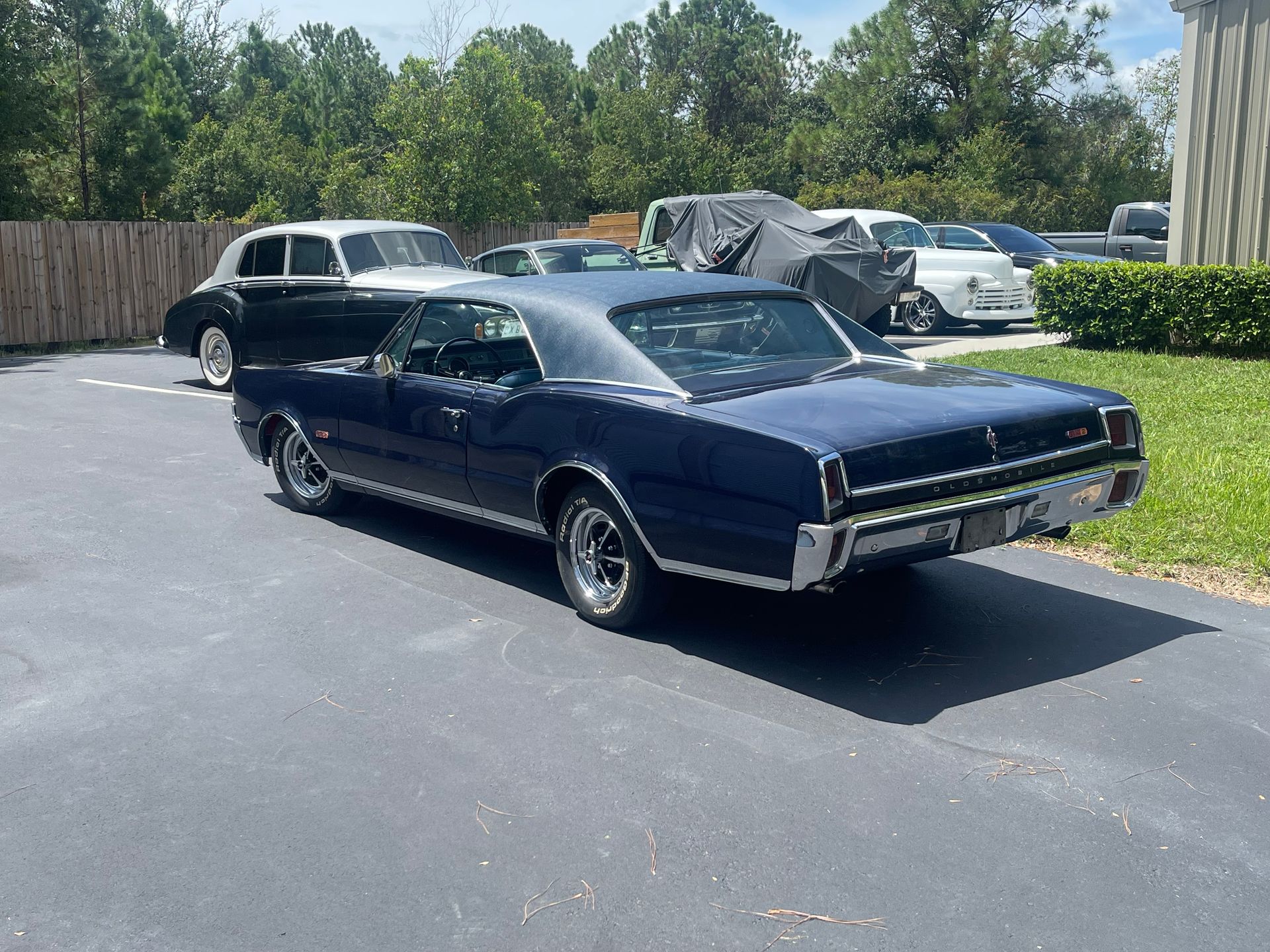 A dark blue classic coupe parked on asphalt next to a silver vintage car in a lot with trees and a building background.