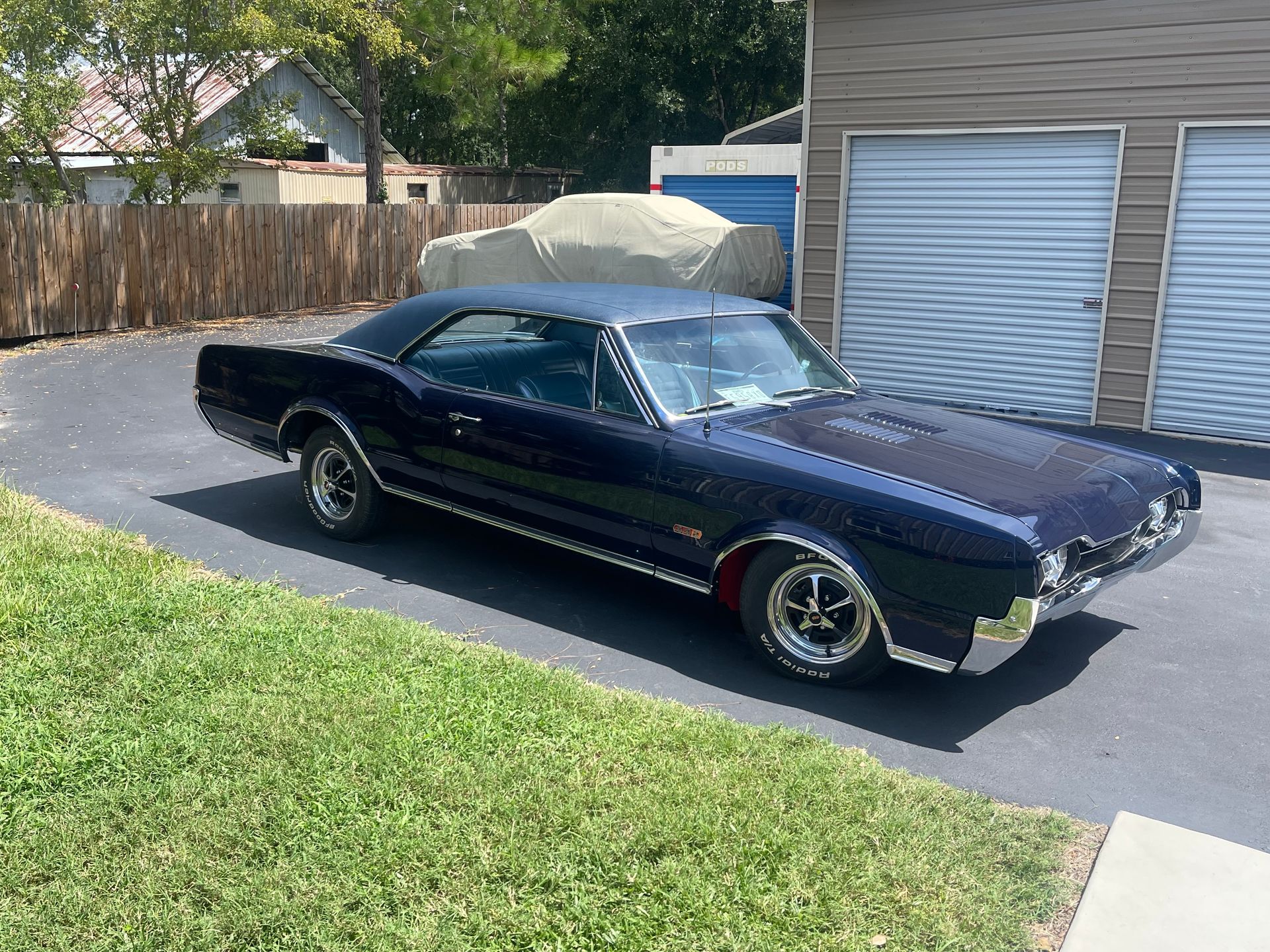 A dark blue vintage muscle car parked on an asphalt driveway next to a tan storage building.