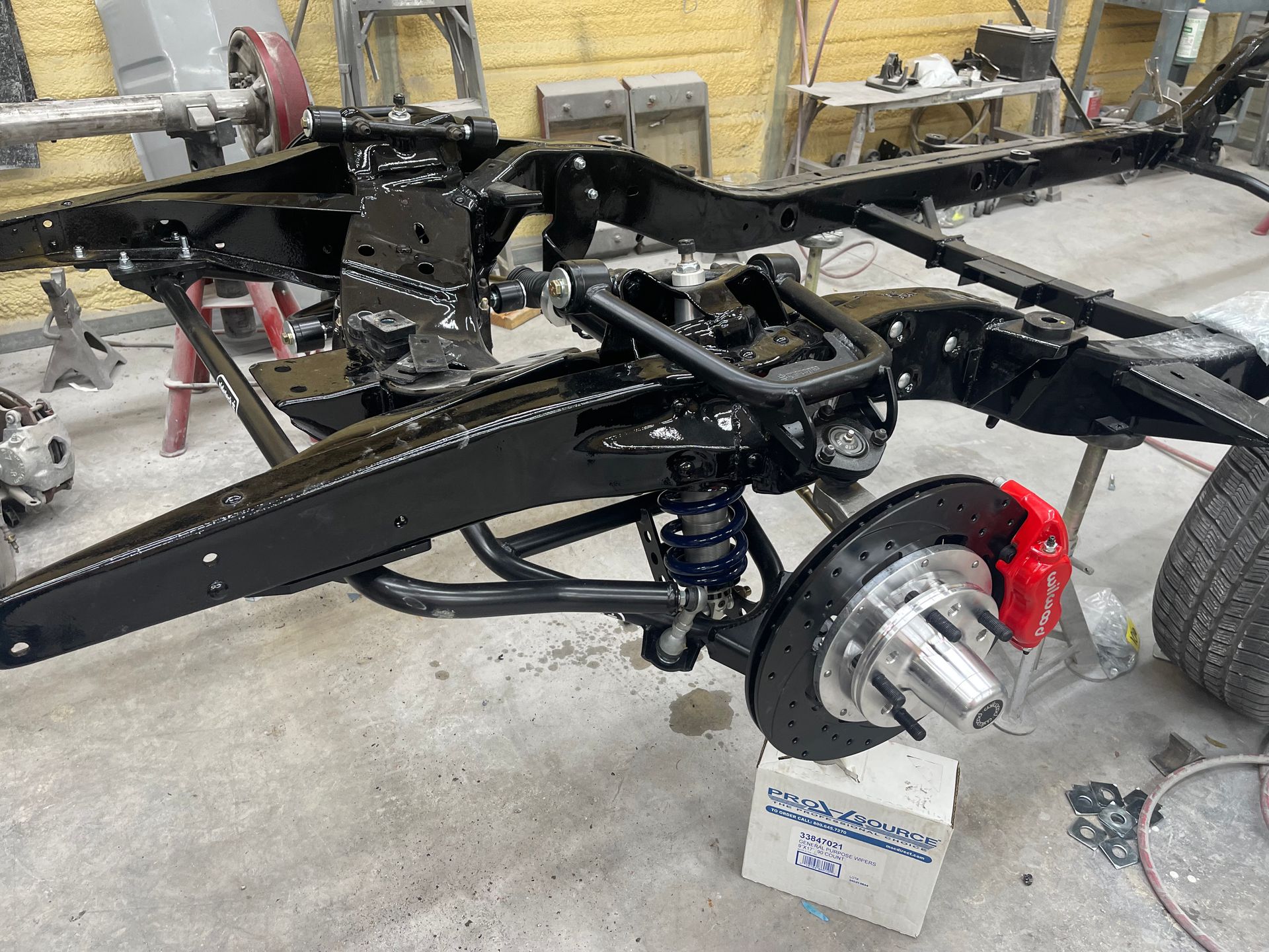 A black vehicle frame with red brake calipers and a modern suspension system, shown in a garage setting.