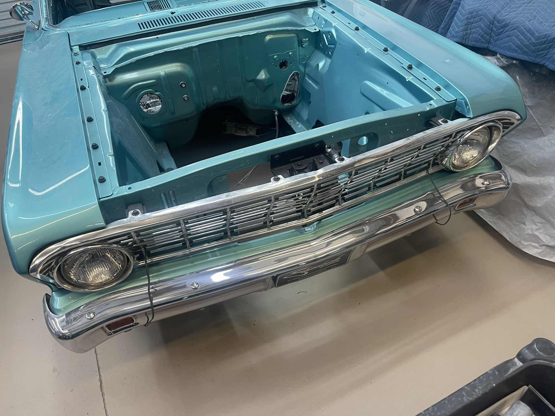 A front-facing view of a light blue classic car body under restoration, featuring a chrome grille and headlight trim.