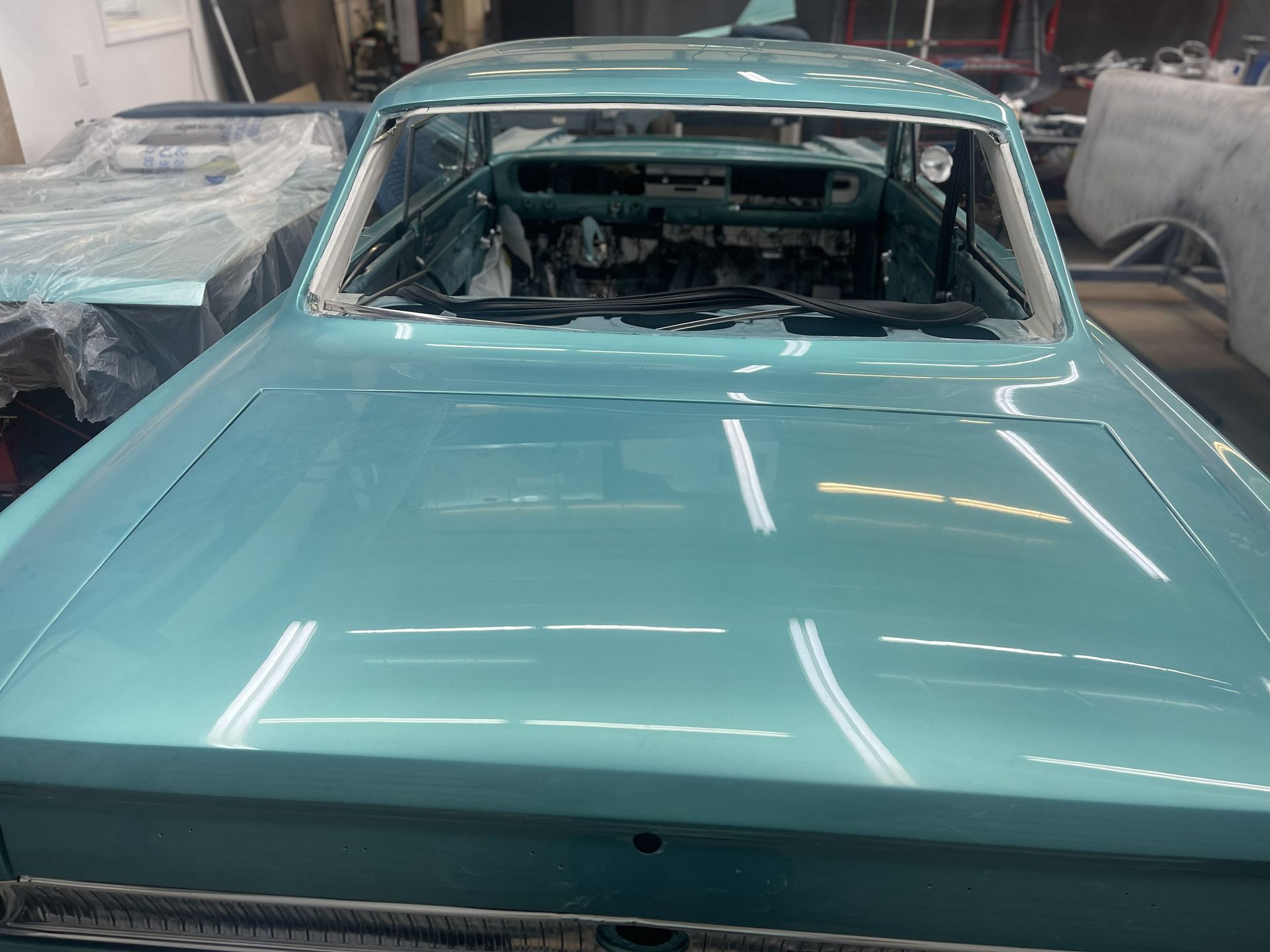 A glossy, light blue classic car body inside an auto body shop, with the interior visible through the windshield.