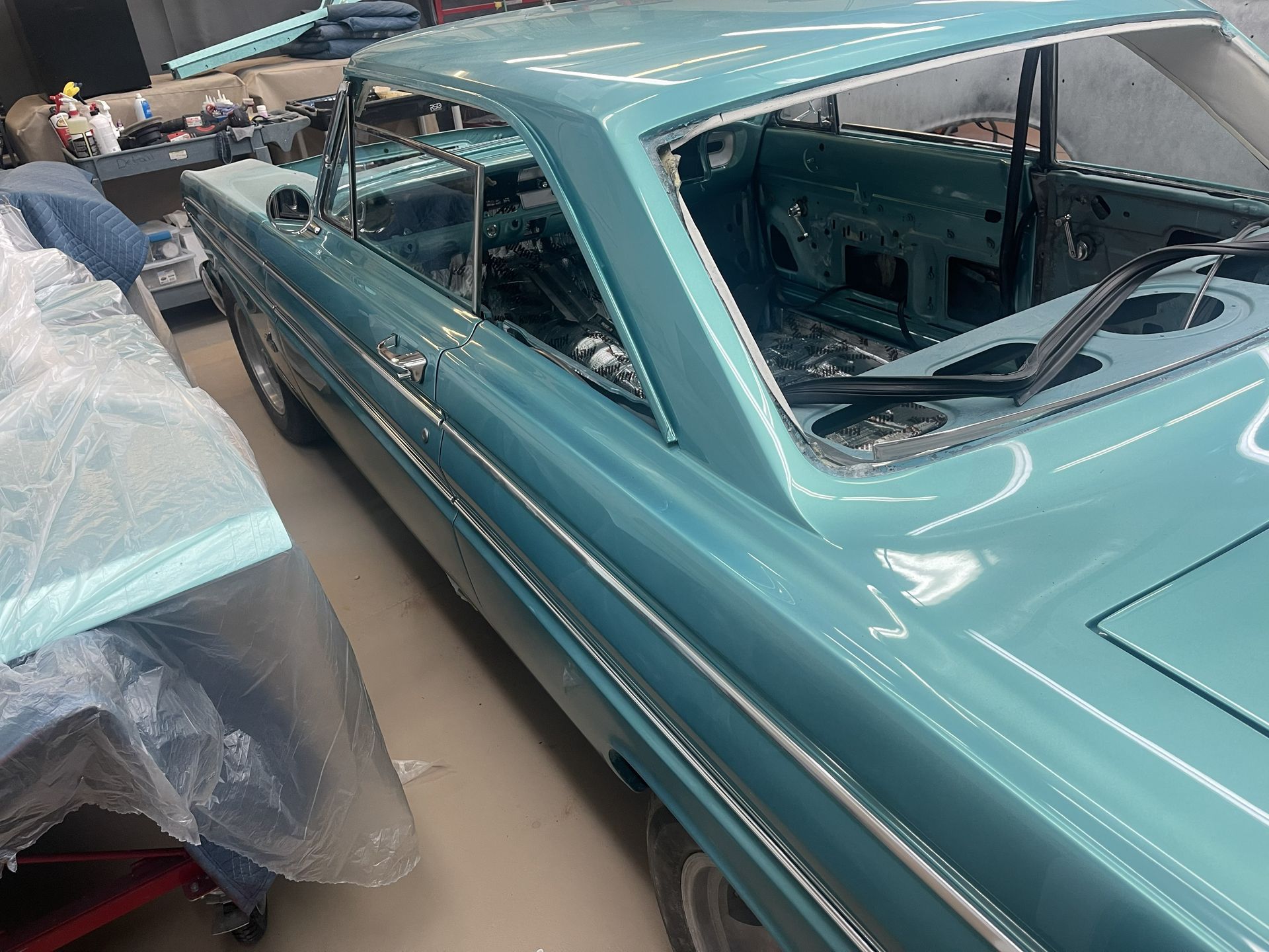 A turquoise classic car body being restored in a workshop, featuring chrome side trim and an empty interior.