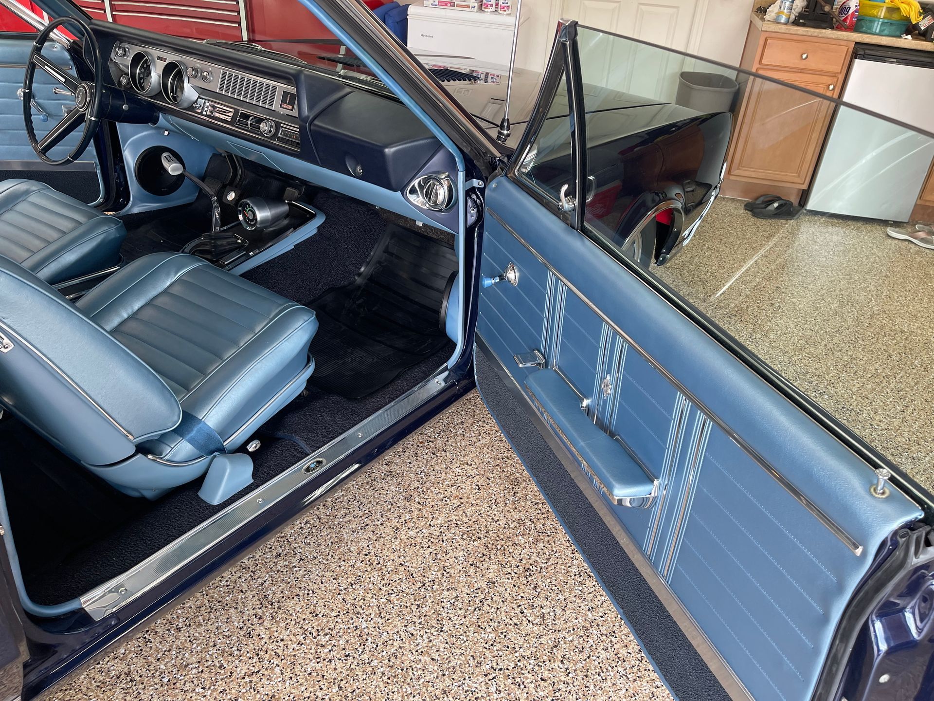 The interior of a classic blue car with vinyl bucket seats, a dashboard with chrome trim, and an open driver-side door.