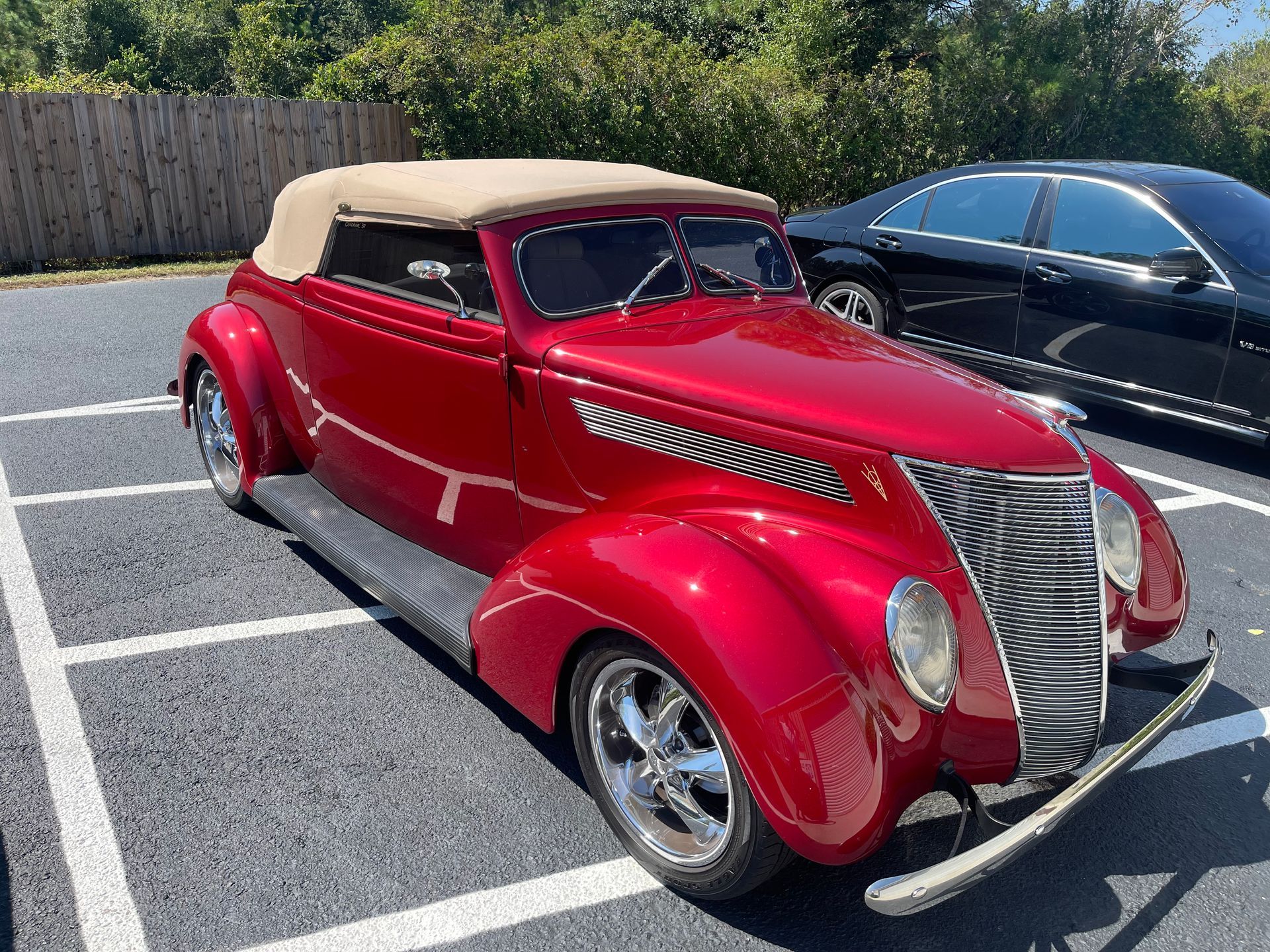 A shiny cherry-red vintage convertible car with a beige soft top, parked in a sunny outdoor parking lot.