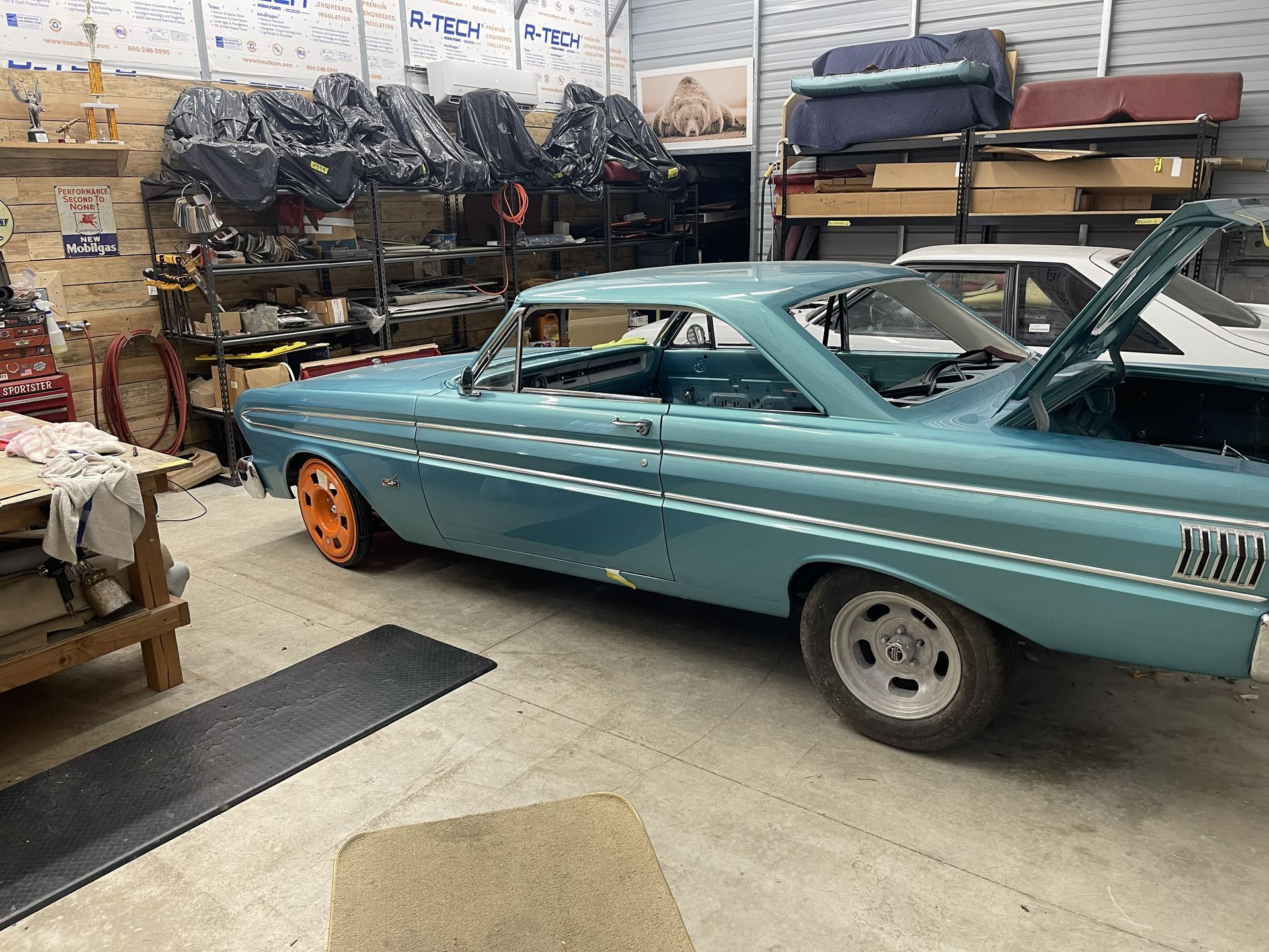 A light blue classic muscle car parked in a garage with mismatched orange and silver wheels and an open trunk.