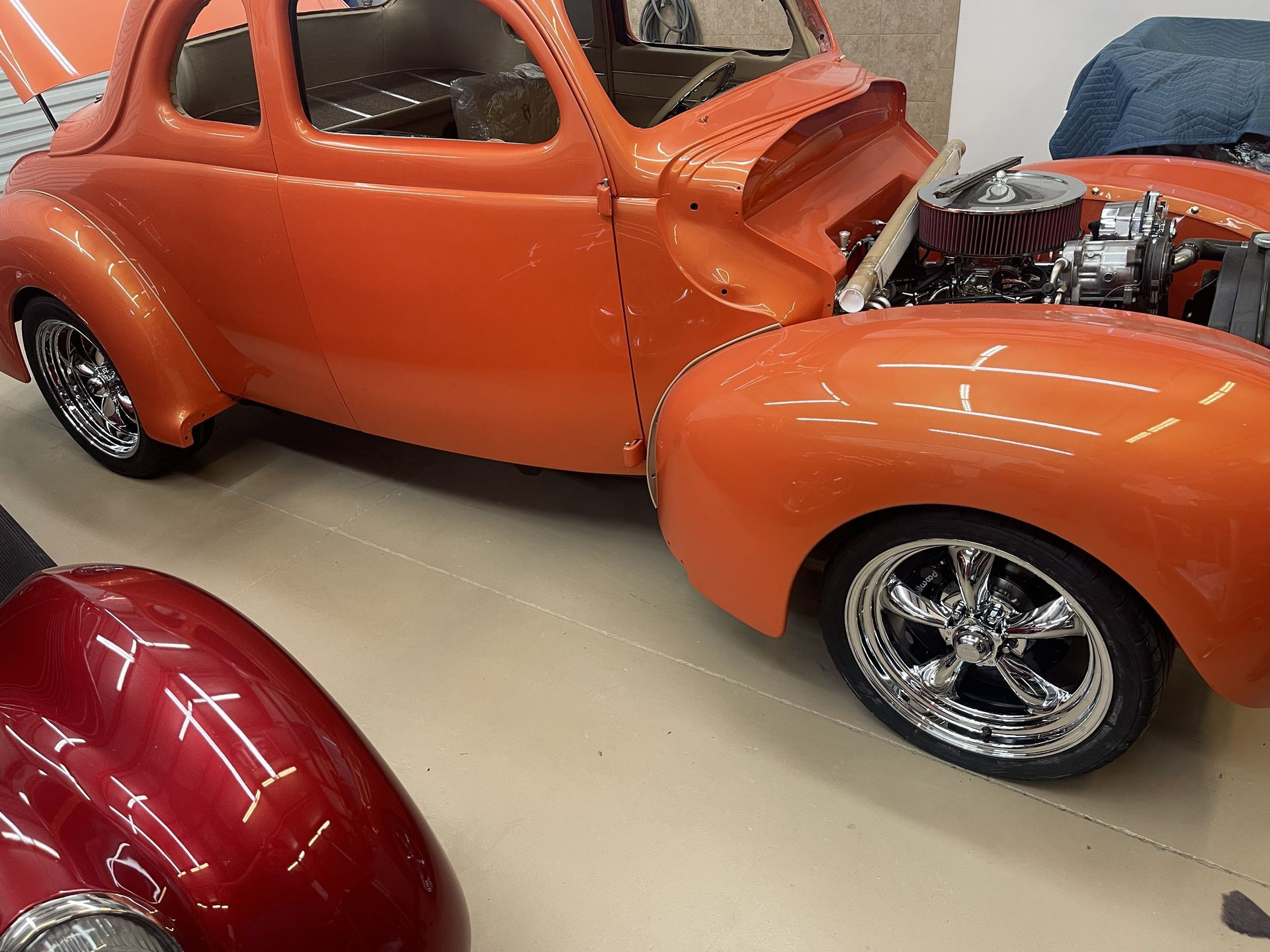 A classic orange vintage car with chrome wheels sits in a garage next to a portion of a shiny red car.