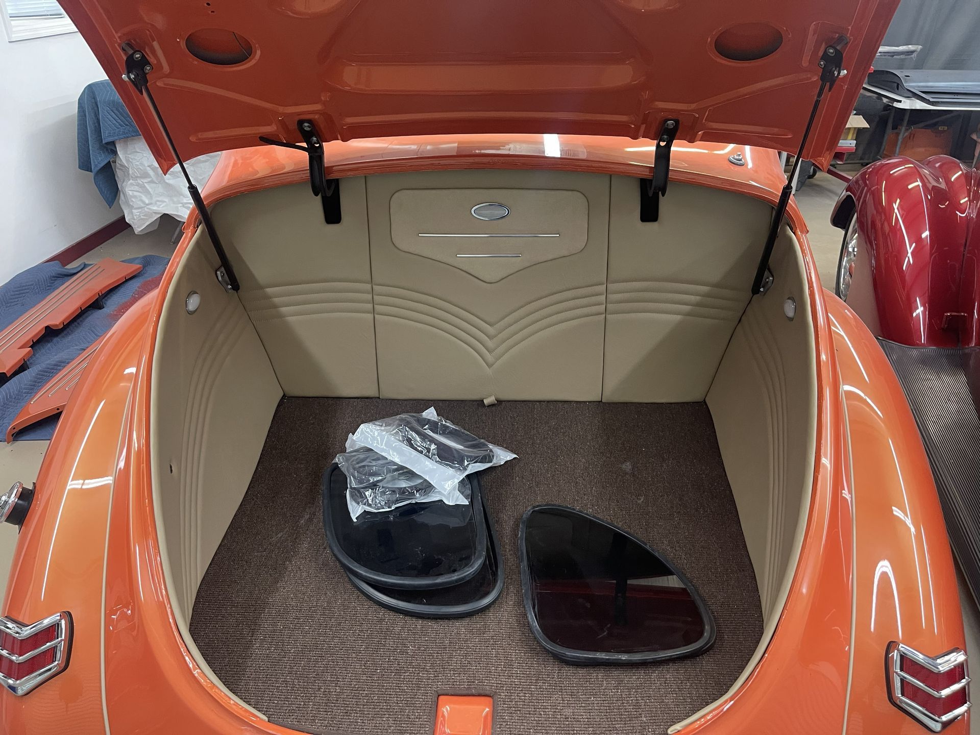 Open trunk of a bright orange classic car with tan custom interior paneling and dark floor mats, inside a garage.