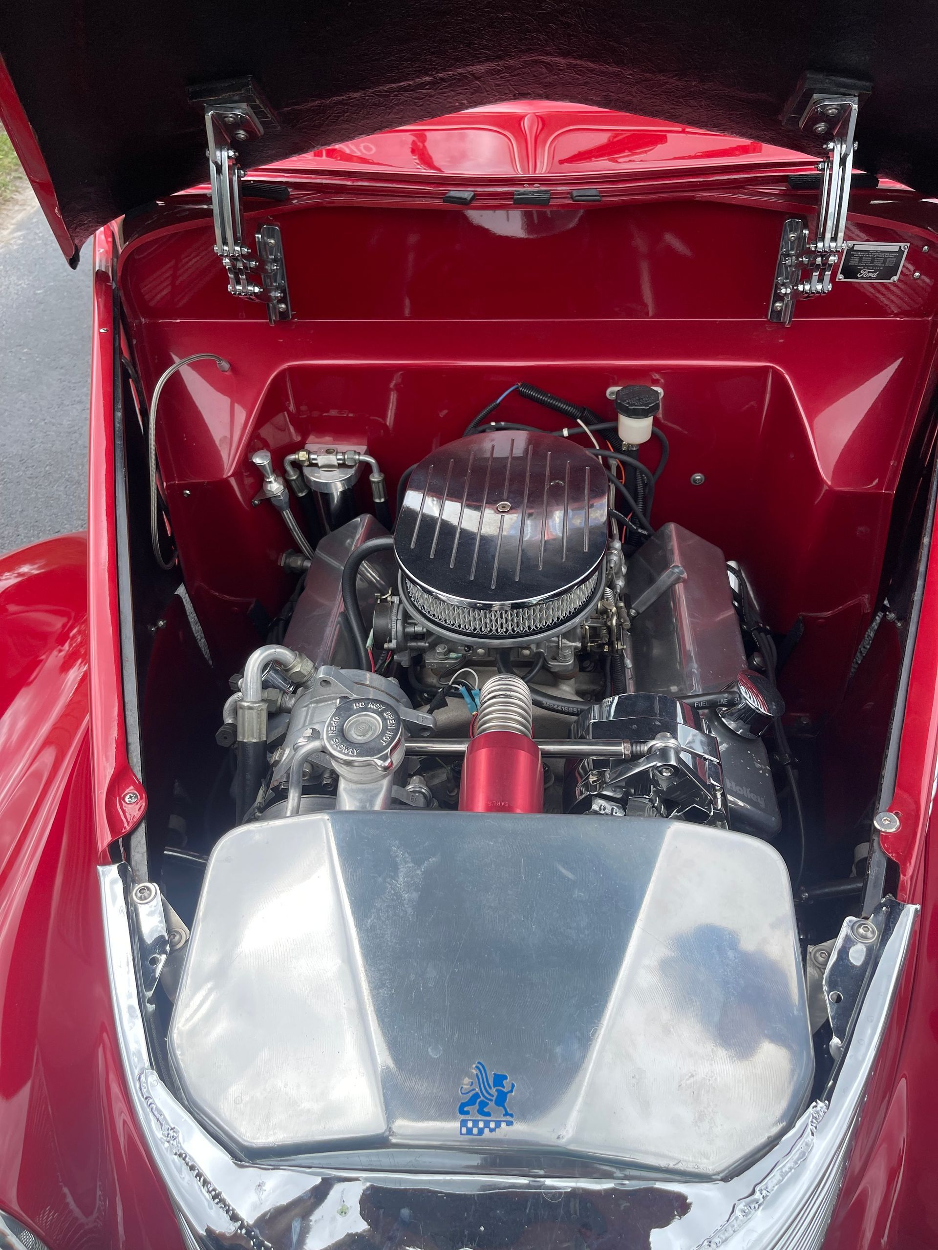 The open hood of a bright red vintage car reveals a detailed engine bay with a polished radiator cover and black engine.