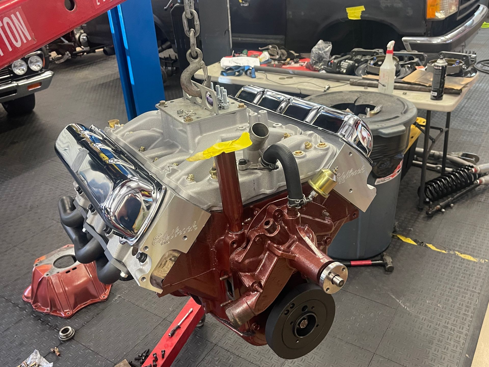 A partially assembled V8 engine suspended from a shop hoist in a garage setting with chrome valve covers.