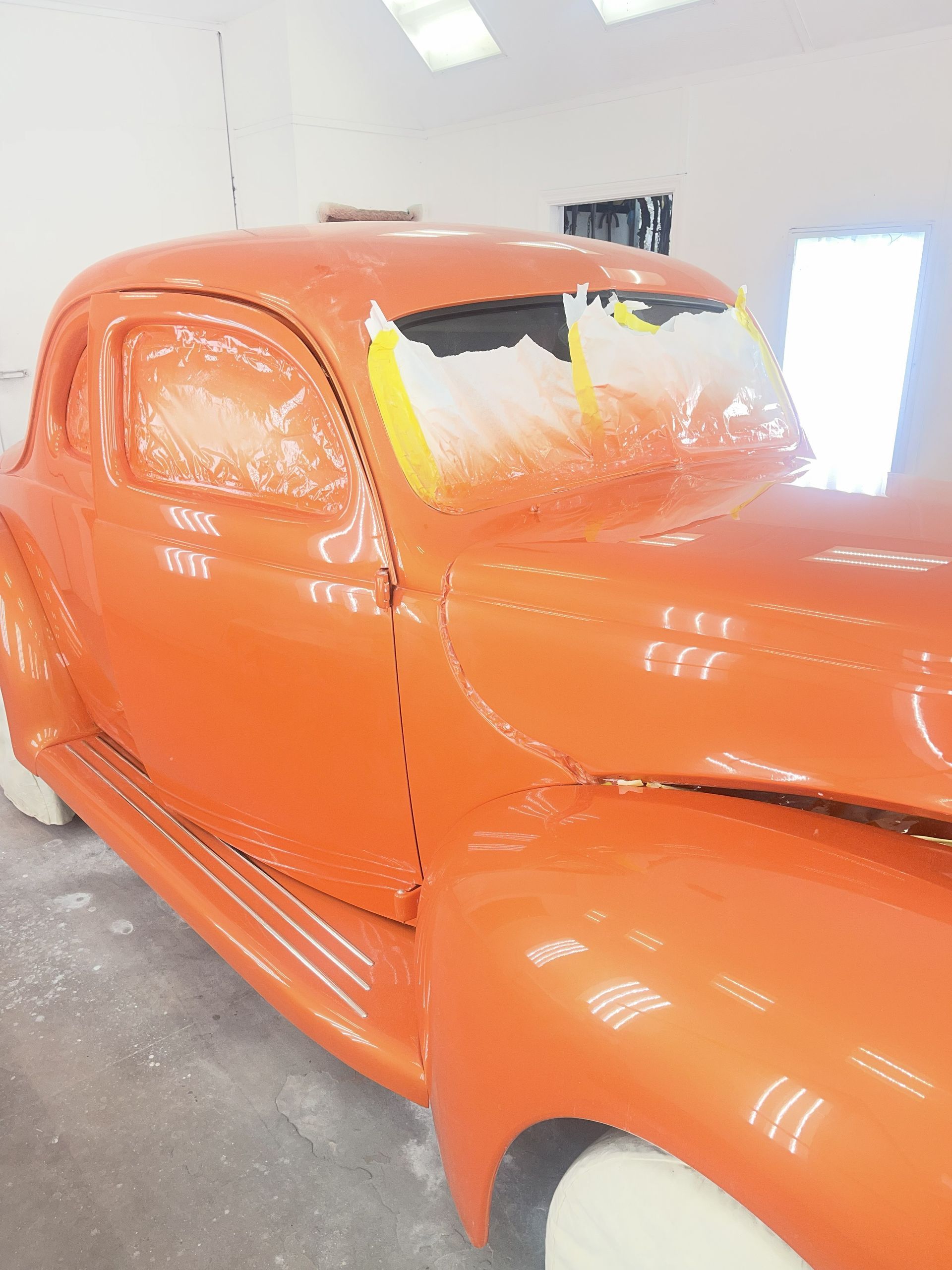 A bright orange vintage car parked in a garage, with the windshield partially masked for painting.